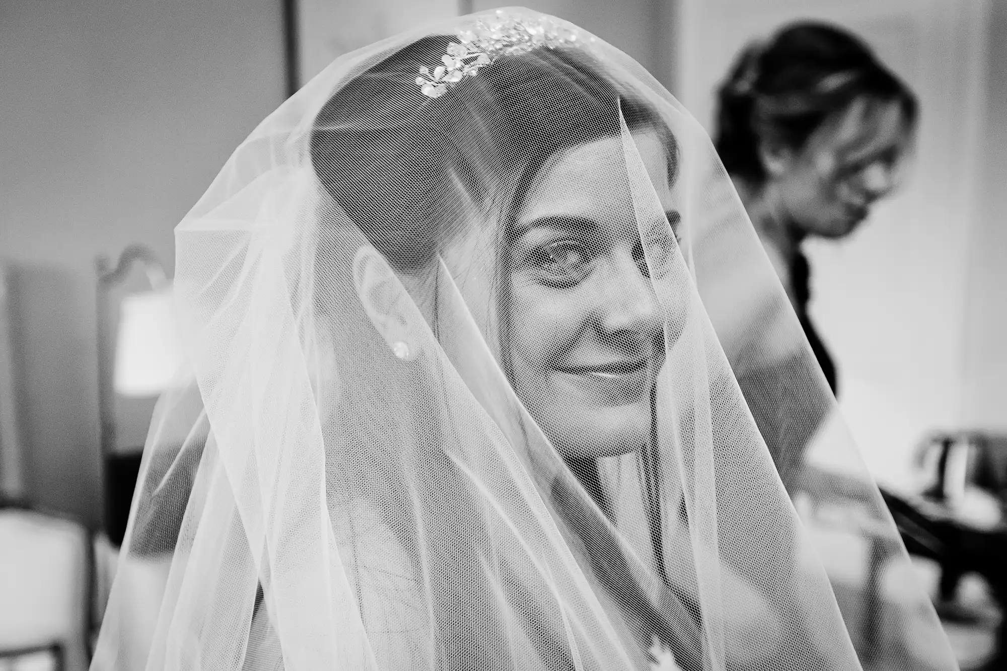 A black and white photo of a smiling bride with a veil over her face, and another woman in the background.