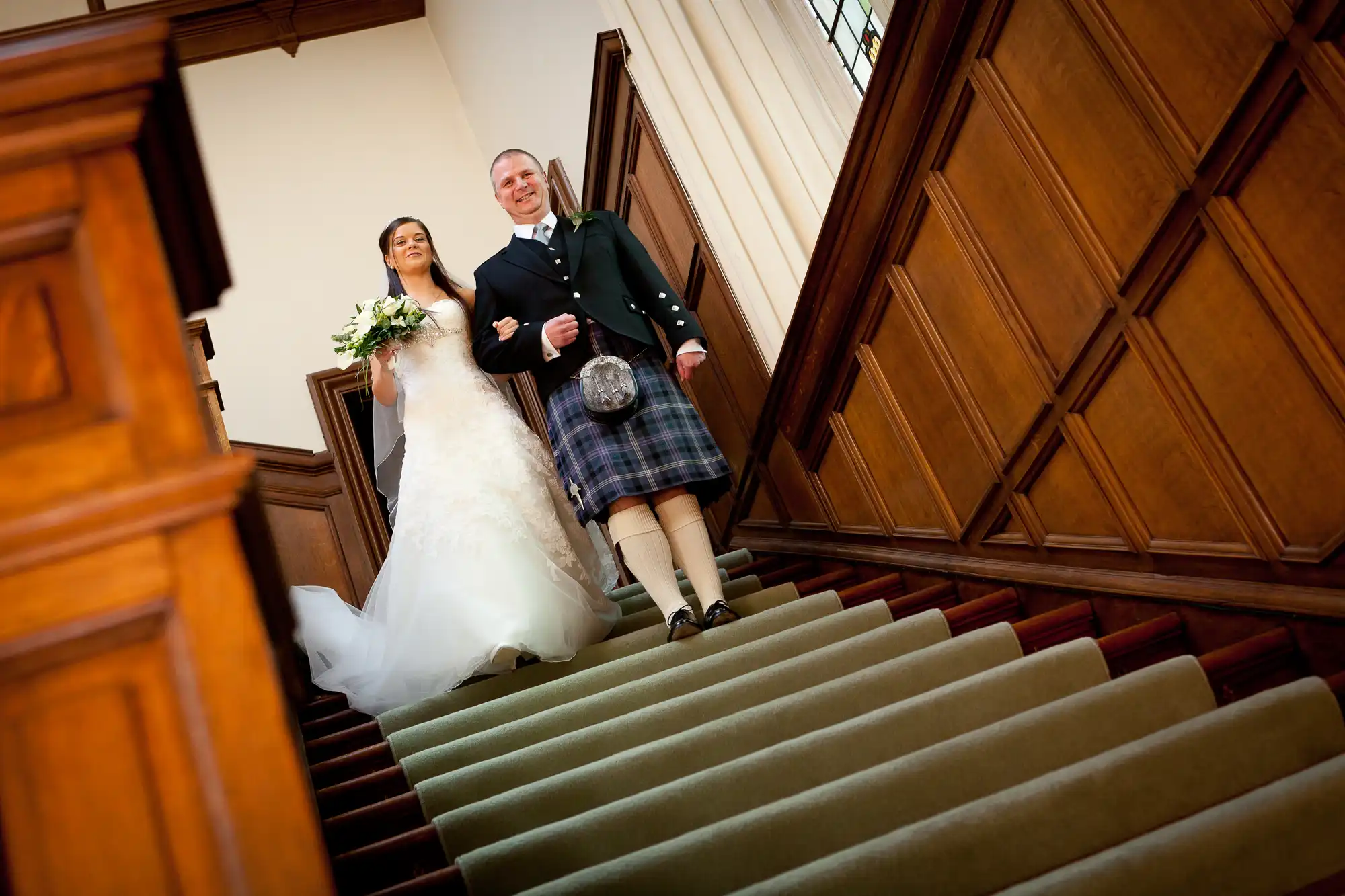 A bride and groom smiling and holding hands as they walk down the stairs of a church, the groom wearing a kilt.
