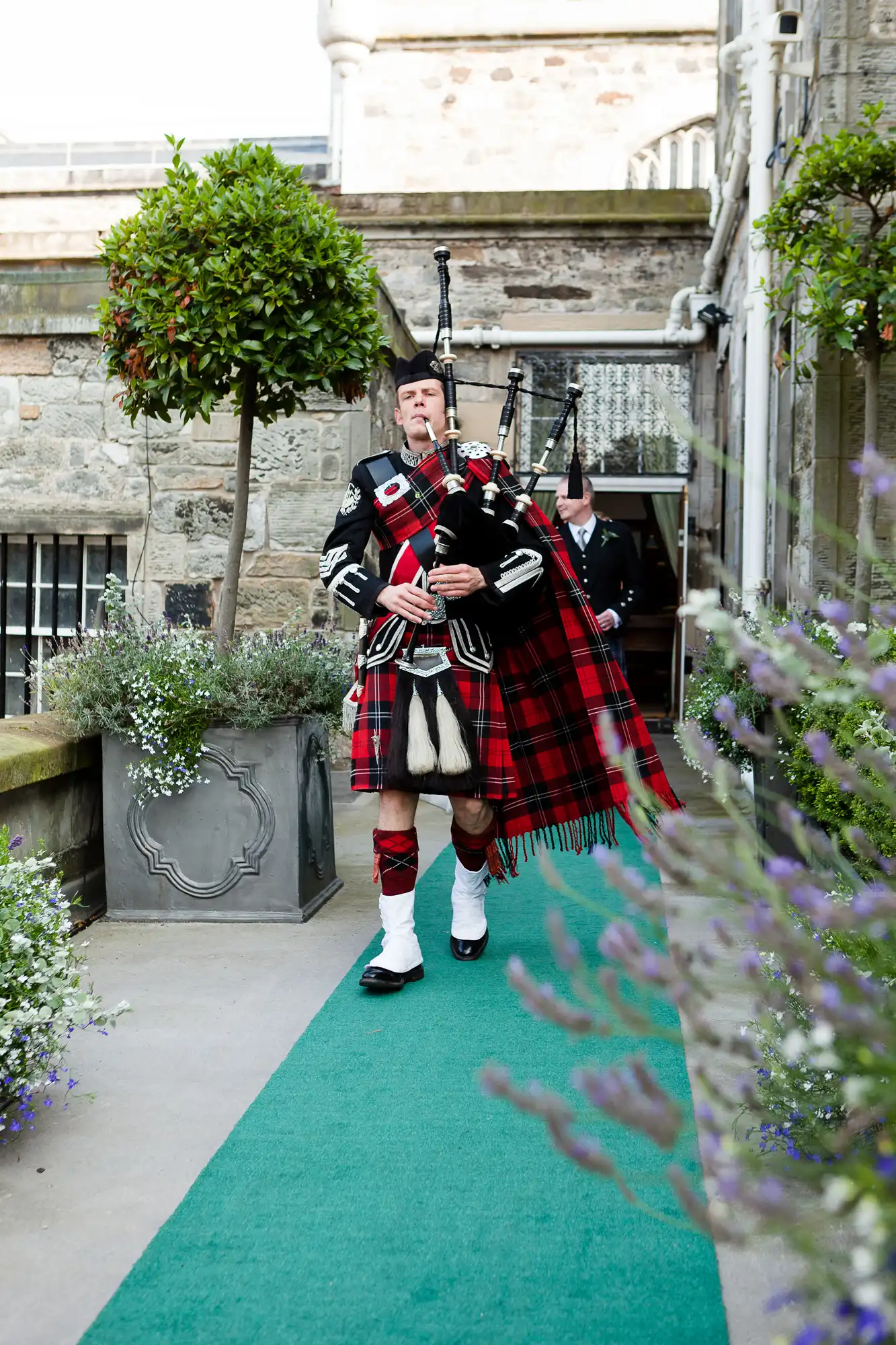A bagpiper in traditional scottish attire, including a tartan kilt, playing bagpipes in a courtyard with topiary plants.