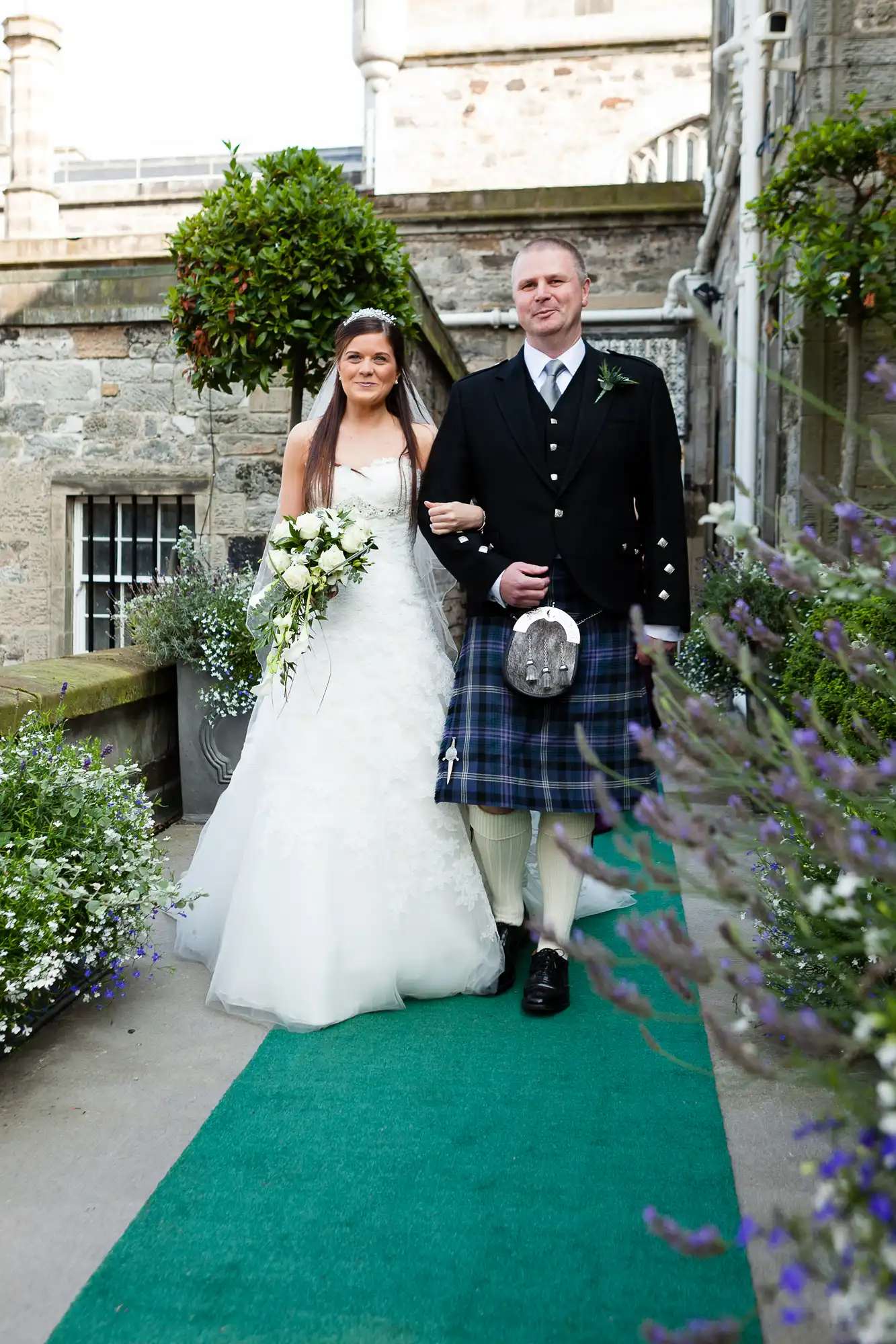 Bride in a white dress and groom in a kilt standing on a green carpet surrounded by flowers at a castle wedding.