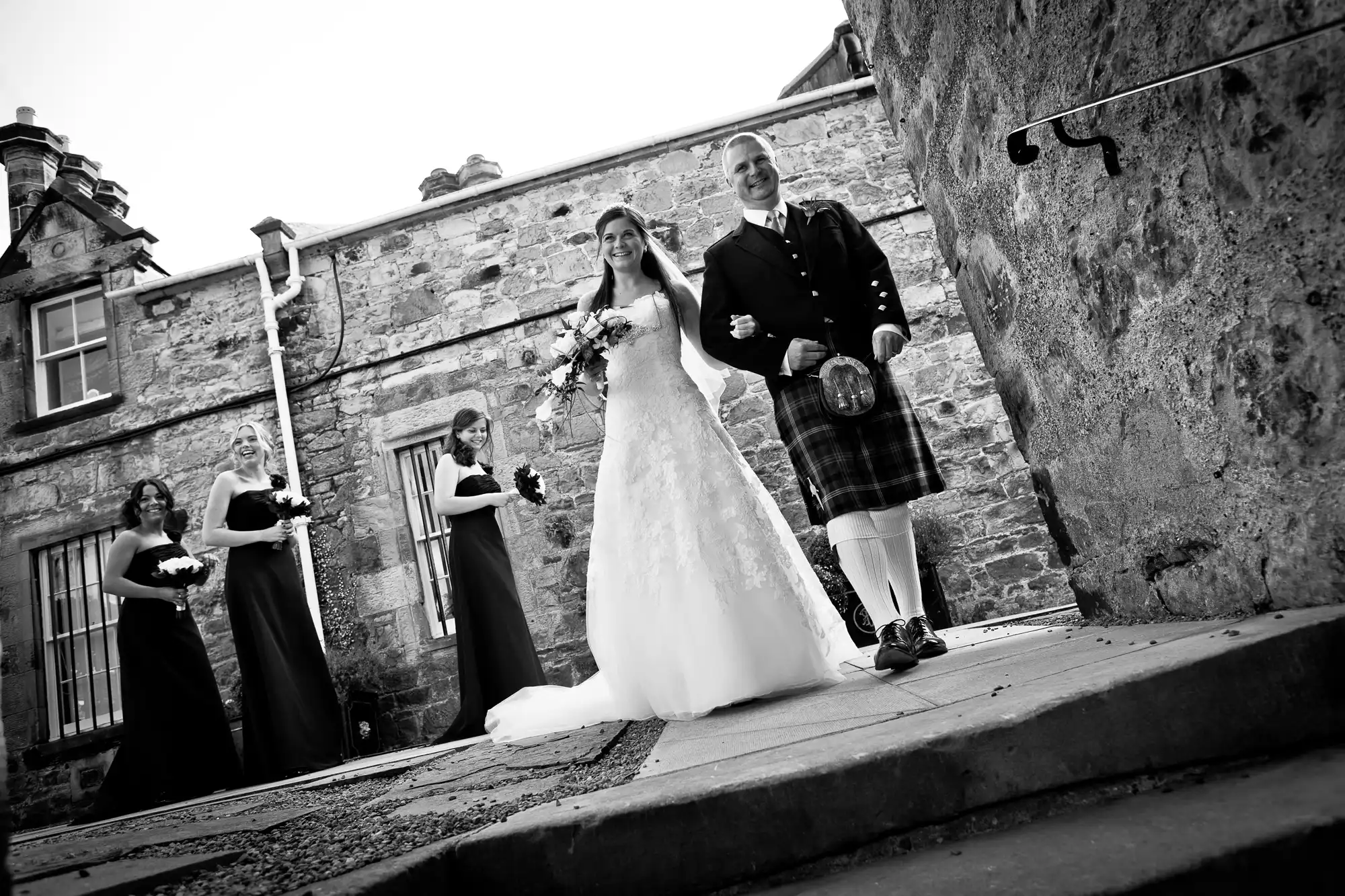 Bride and groom walking happily, the groom in a kilt, accompanied by bridesmaids in black dresses, outside a stone building.