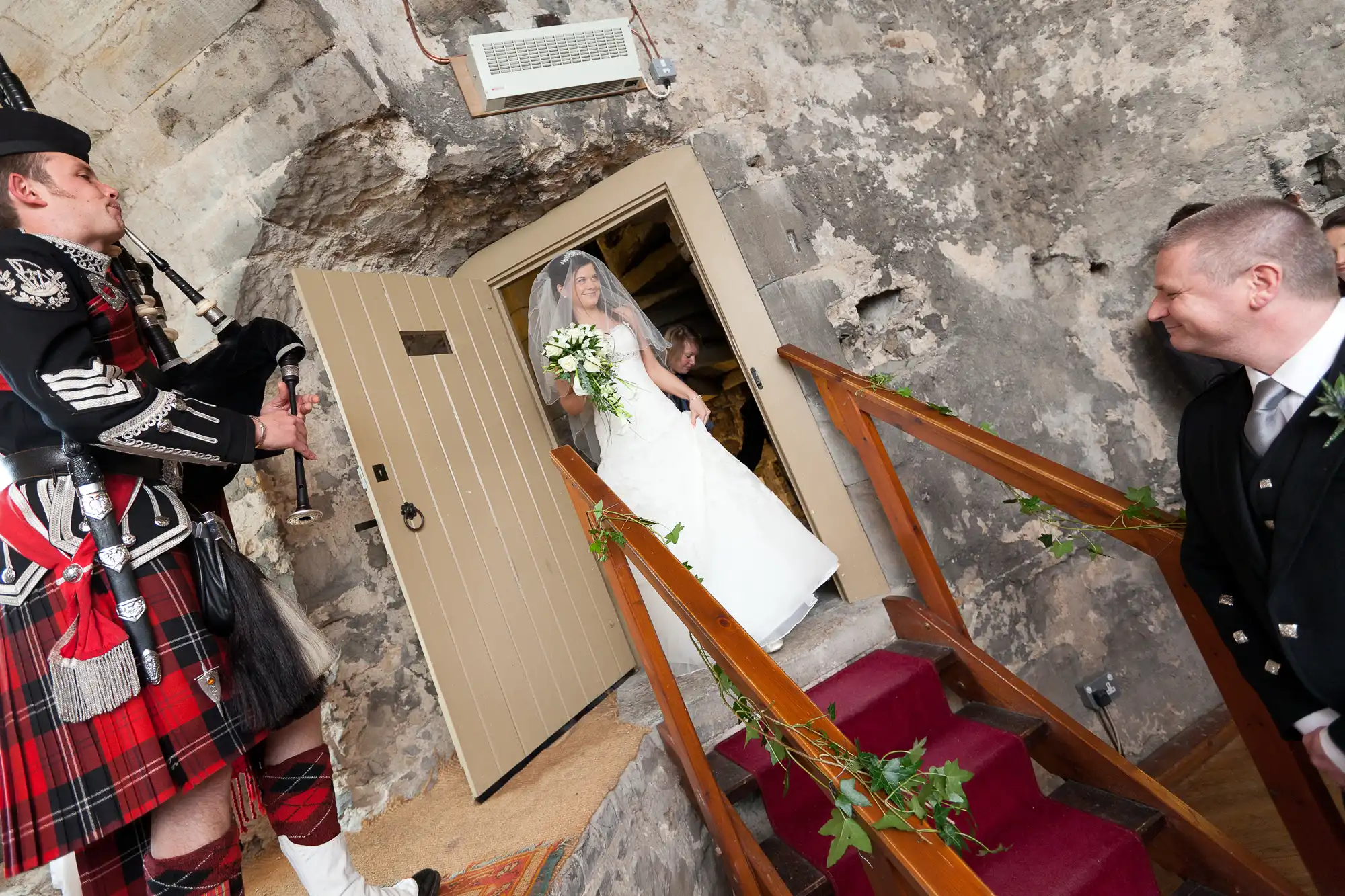 A bride in a white dress holding a bouquet descends stairs at a wedding venue, greeted by a man in a kilt and another man in a suit.