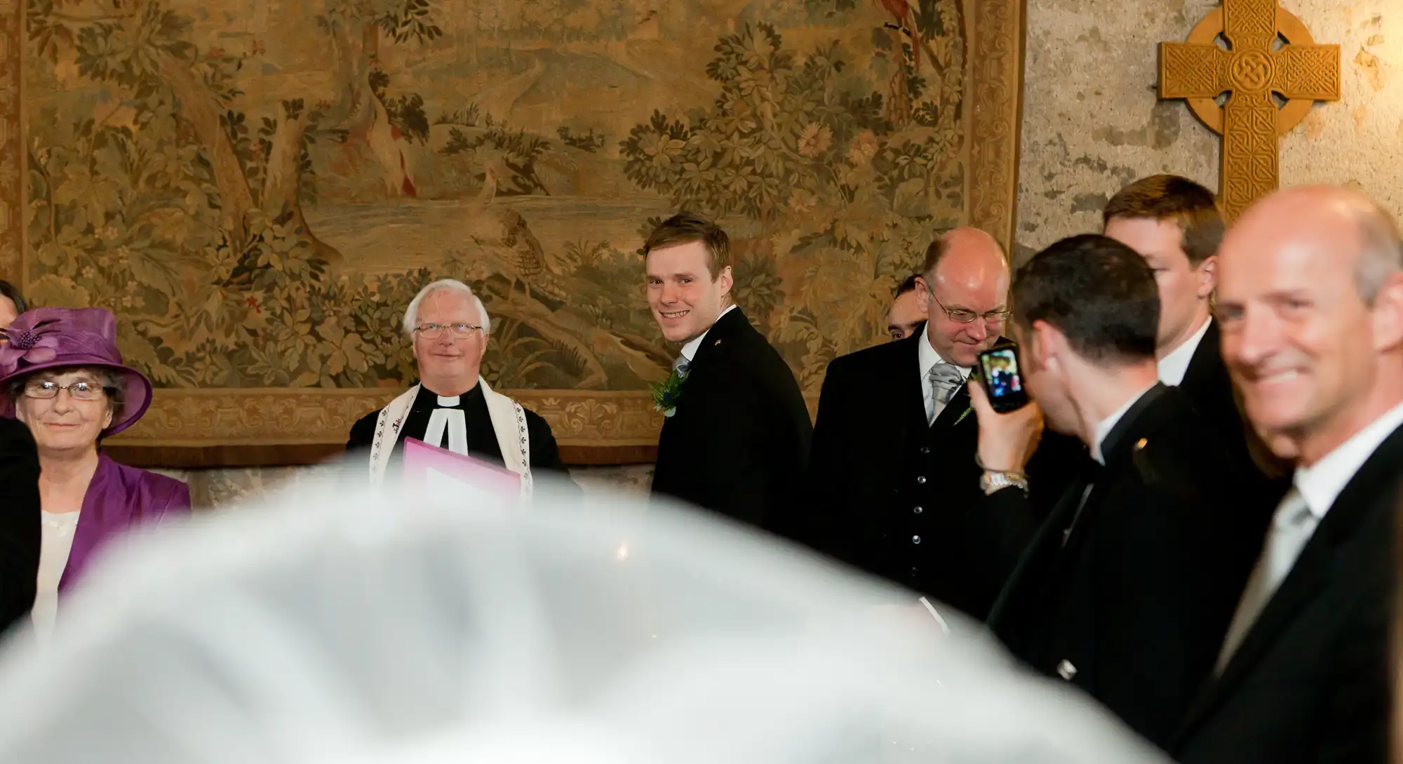 Wedding ceremony in a church with a bride's veil in the foreground and smiling groom looking back, surrounded by guests and a clergyman.