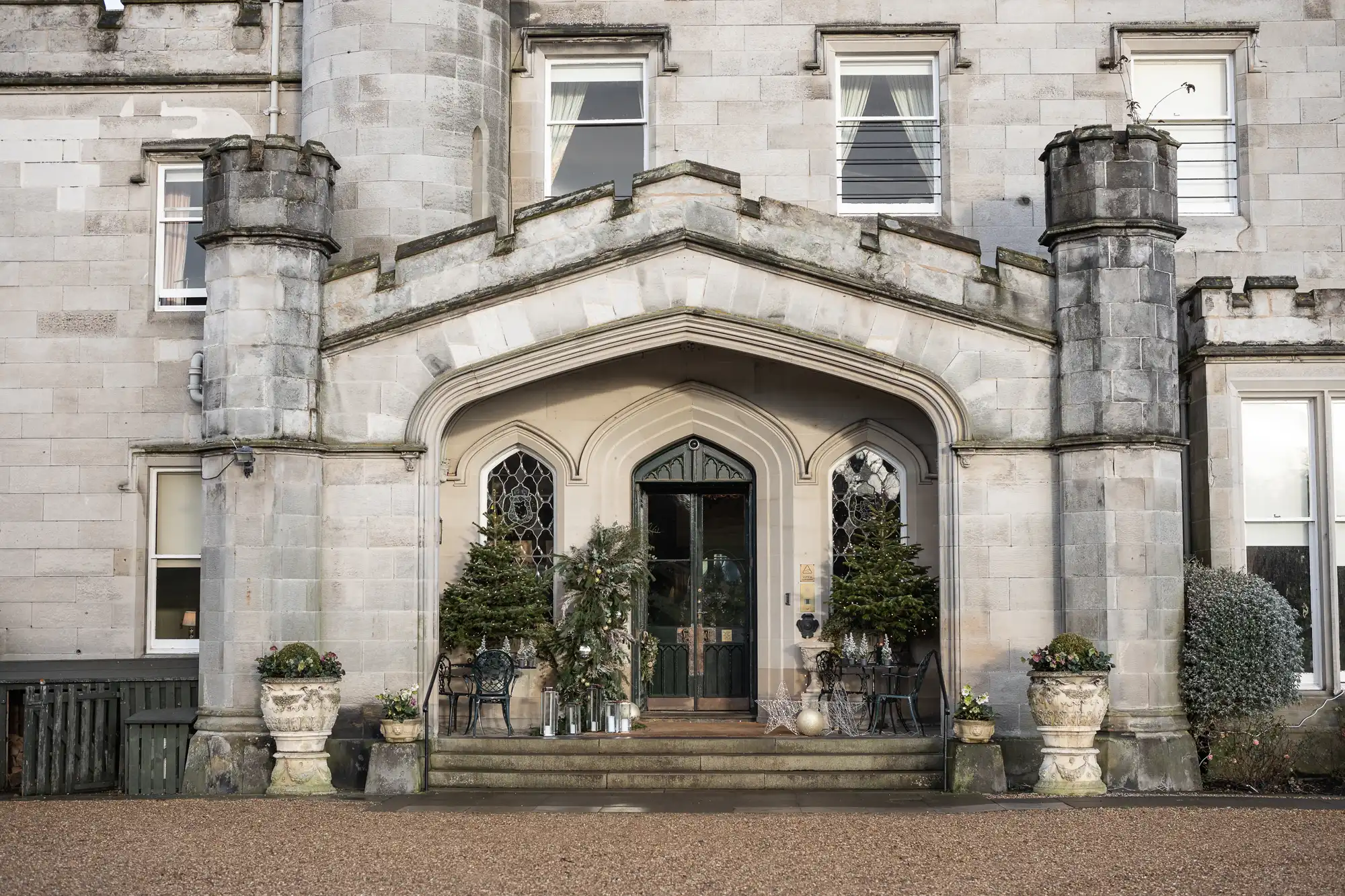 Front entrance of a stone building with a large archway, two cylindrical towers, potted plants, and steps leading to a black door.