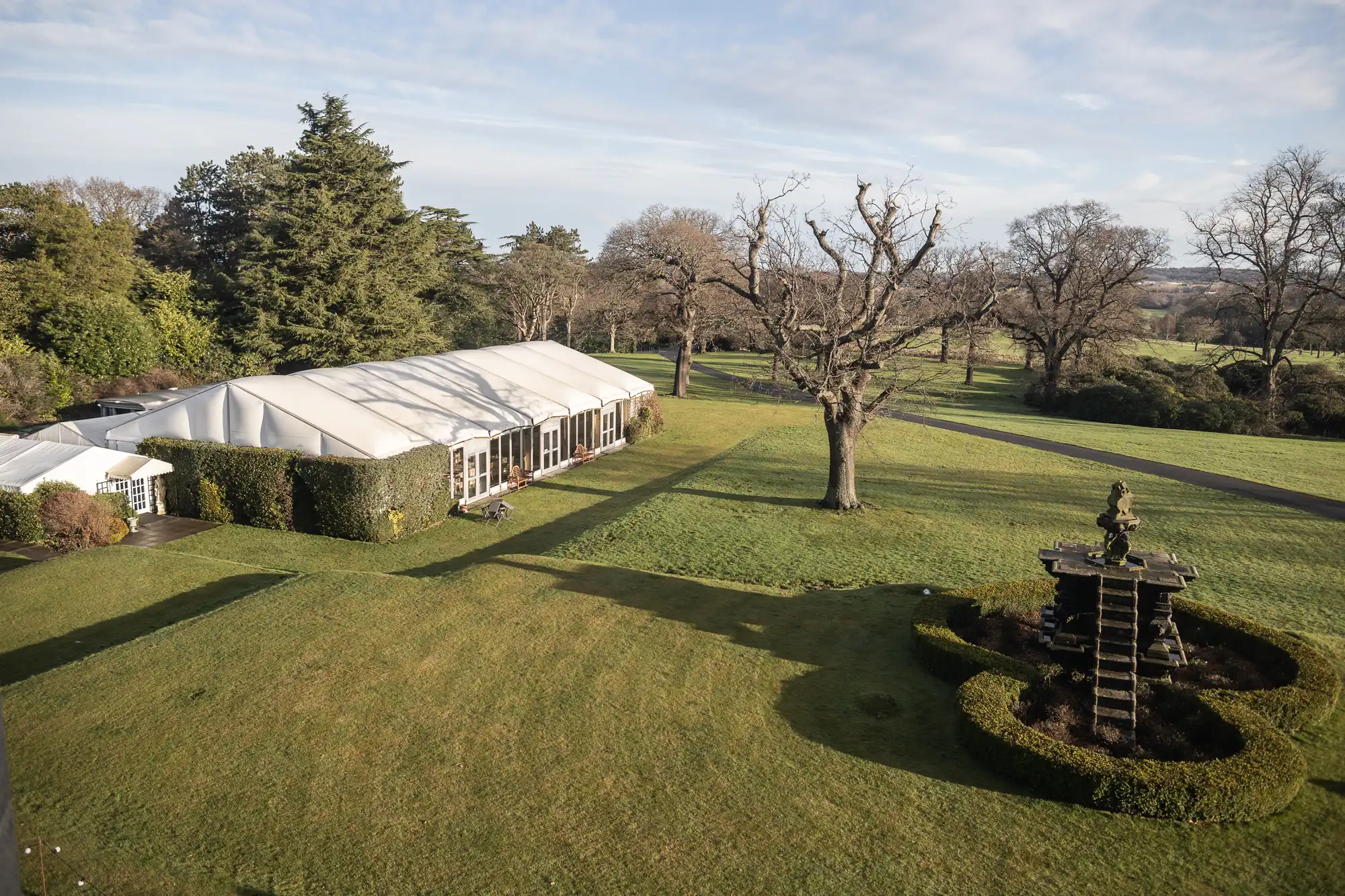 Aerial view of a large white marquee set up on a manicured lawn with hedges, trees, and a fountain in a circular hedge formation in a park-like setting.