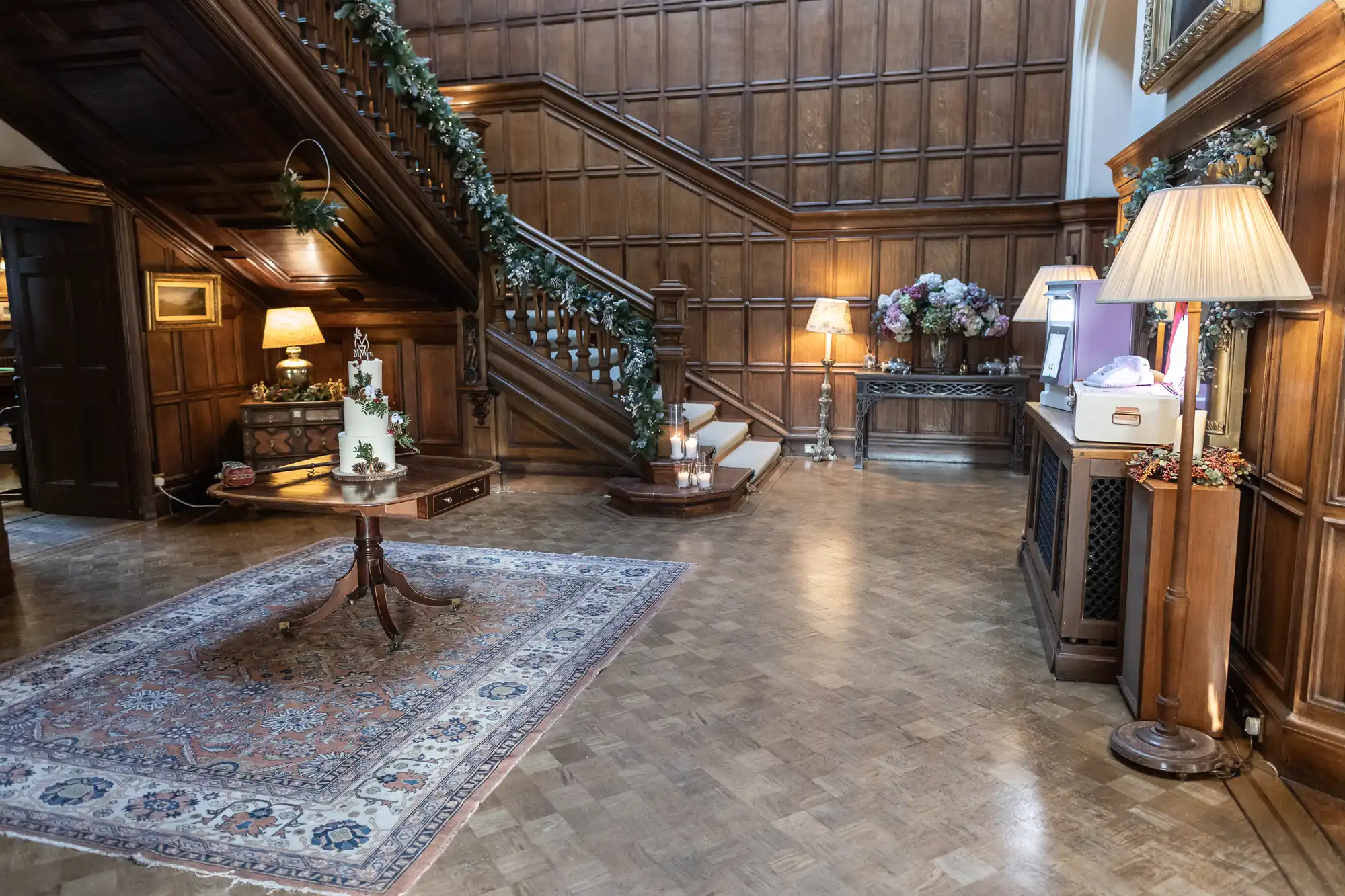 A wood-paneled room with an ornate staircase, a table with decorative items and candles, and a patterned rug. The space is lit by several lamps and decorated with greenery and flowers.