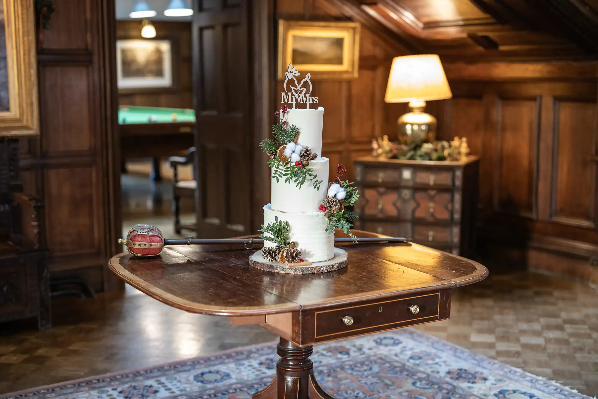 A tiered white cake with a "Mr & Mrs" topper, adorned with festive greenery and pinecones, is displayed on a wooden table. A string instrument and bow lie beside it in a decorated wood-paneled room.