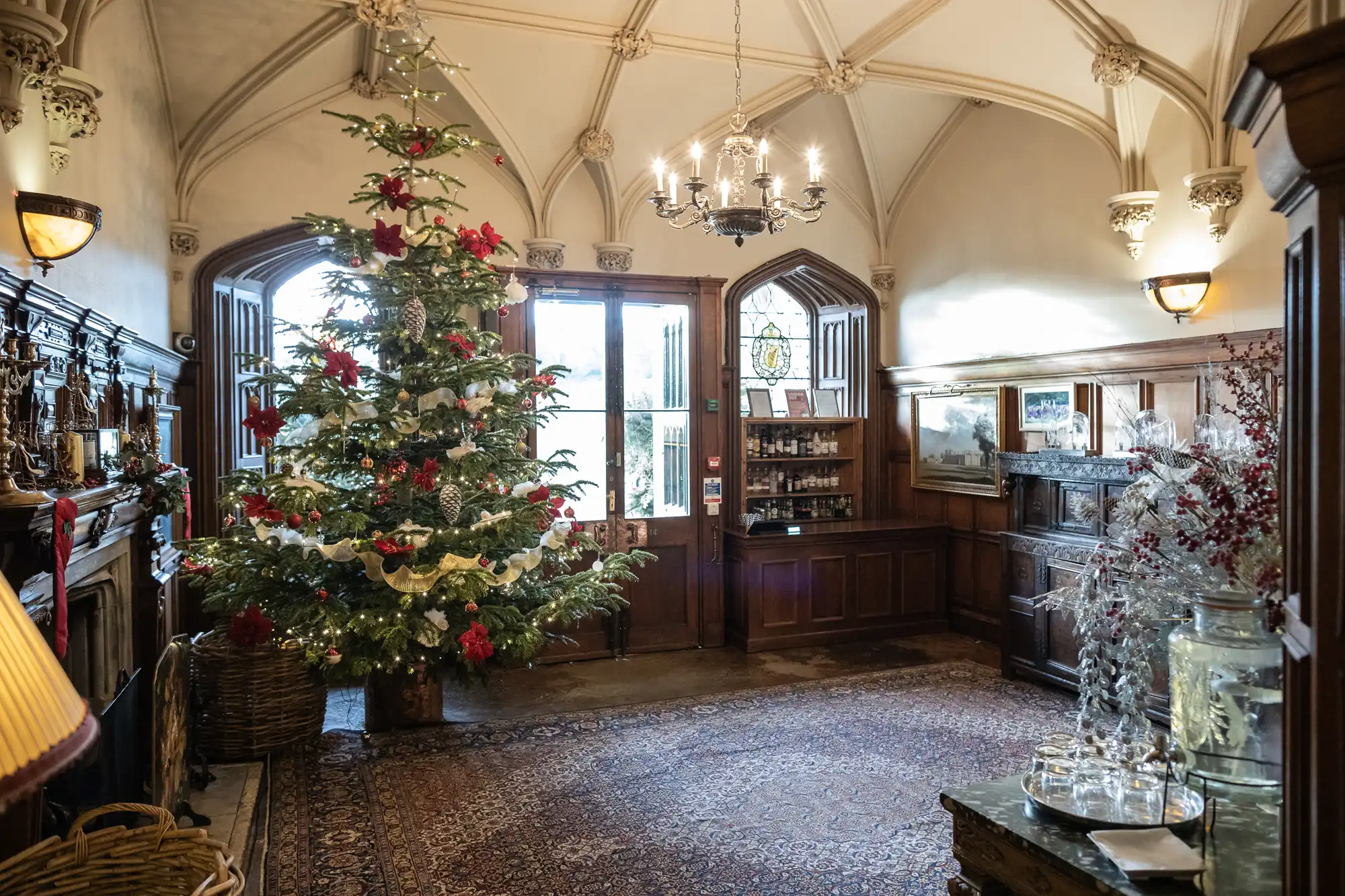 A decorated Christmas tree stands in a grand room with ornate ceiling arches, a chandelier, and wood paneling. The space features a fireplace, patterned rug, and various decorative items.