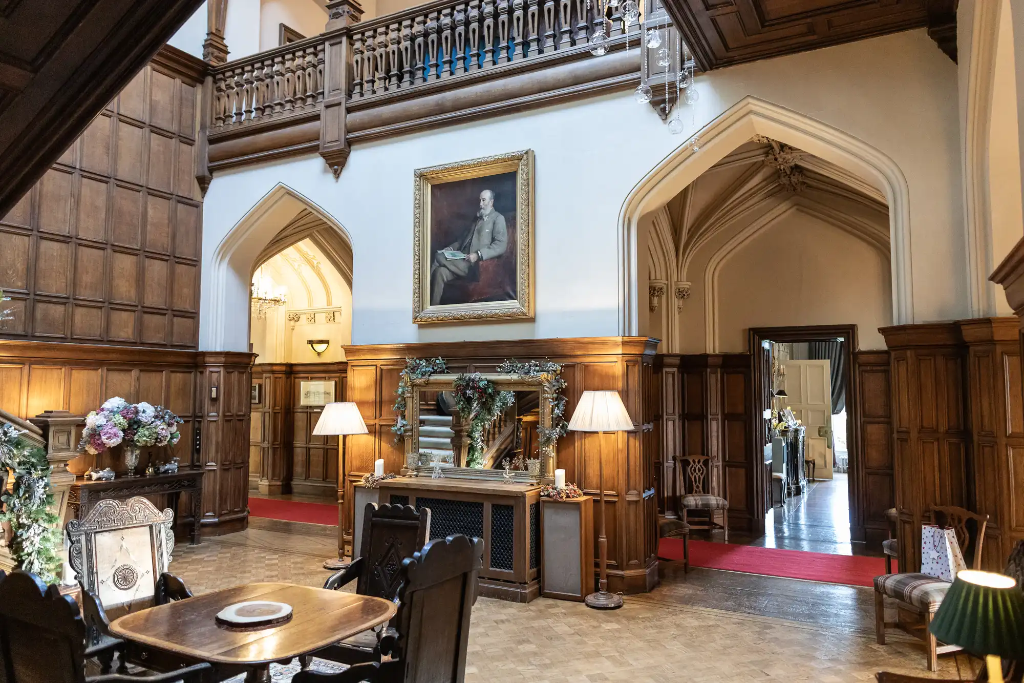 An elegant hall with wood paneling, archways, a central table with chairs, a fireplace adorned with decorations, lamps, and a portrait painting above the fireplace.