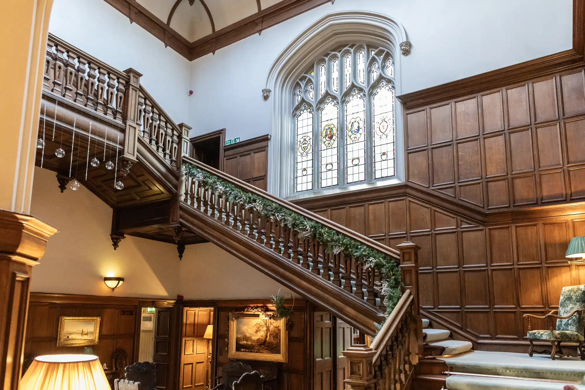 A grand wooden staircase with railings and garland, leading to an upper floor in a building. It features tall, stained glass windows and a partially paneled wall. The lower area has artwork and furniture.