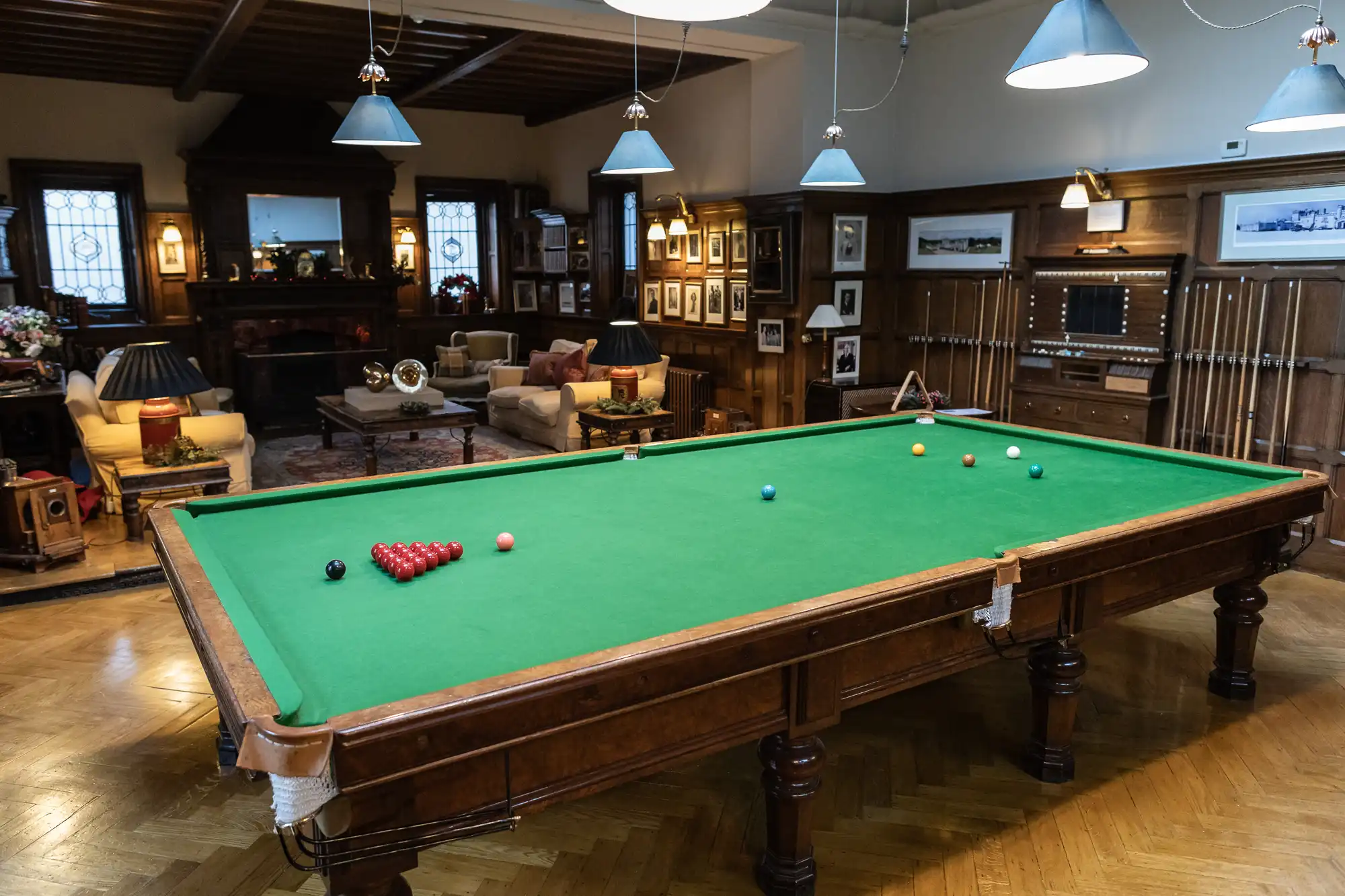 A spacious room featuring a large snooker table in the center. The room is elegantly furnished with sofas, lamps, framed pictures, and wooden wall paneling, under warm lighting from hanging lamps.