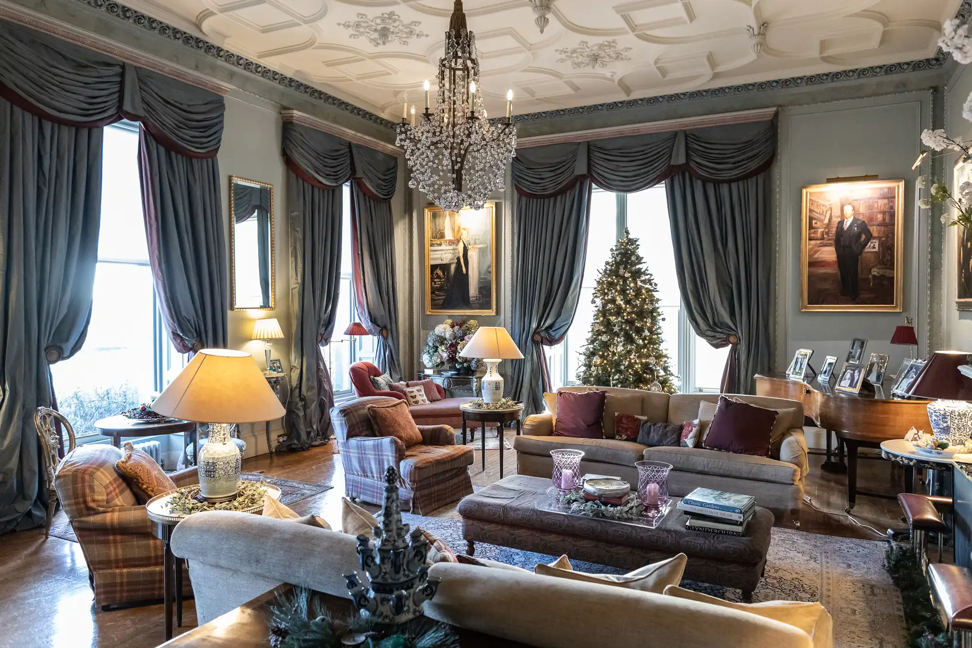 A grand, ornate living room with plush furniture, a chandelier, large draped windows, a Christmas tree, framed portraits, a piano, and lamps providing warm light.