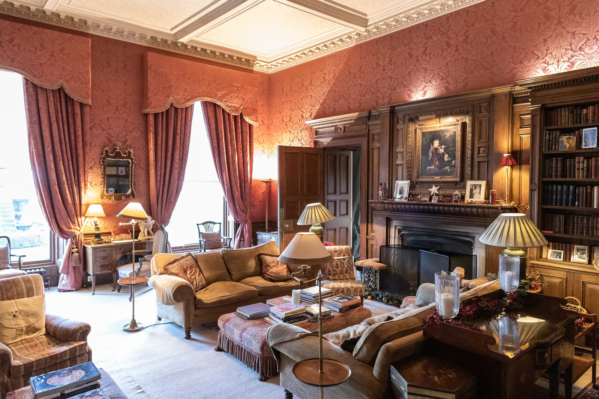 A cozy, traditional living room features ornate red wallpaper, framed artwork above a fireplace, plush sofas, and well-stocked bookshelves, with floor lamps providing warm lighting.