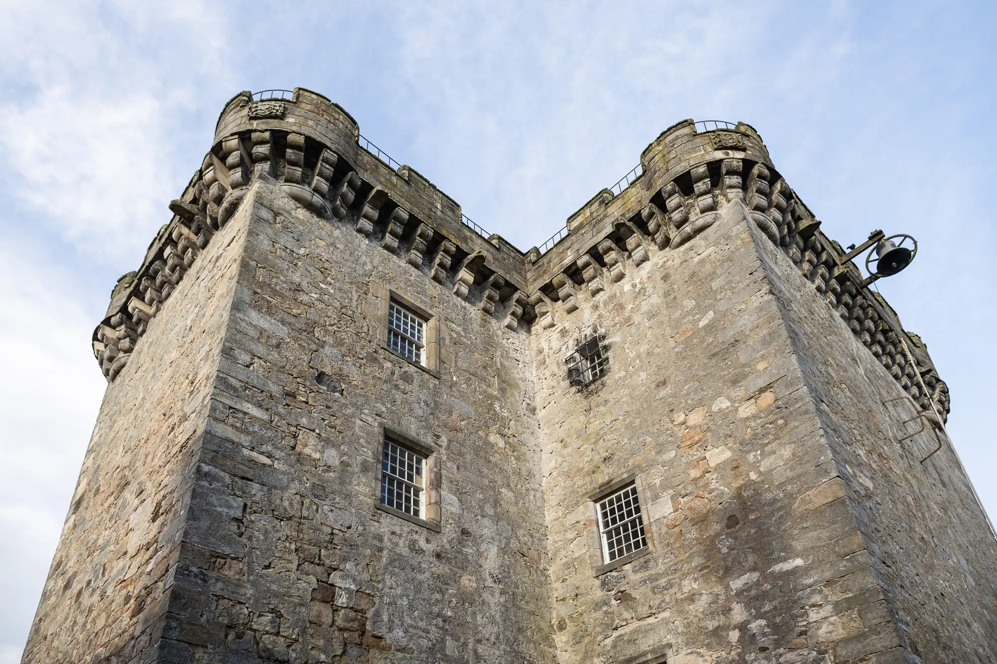 A close-up view of a tall, stone castle tower with small windows, battlements at the top, and a bell on the right side. The sky is partially cloudy in the background.