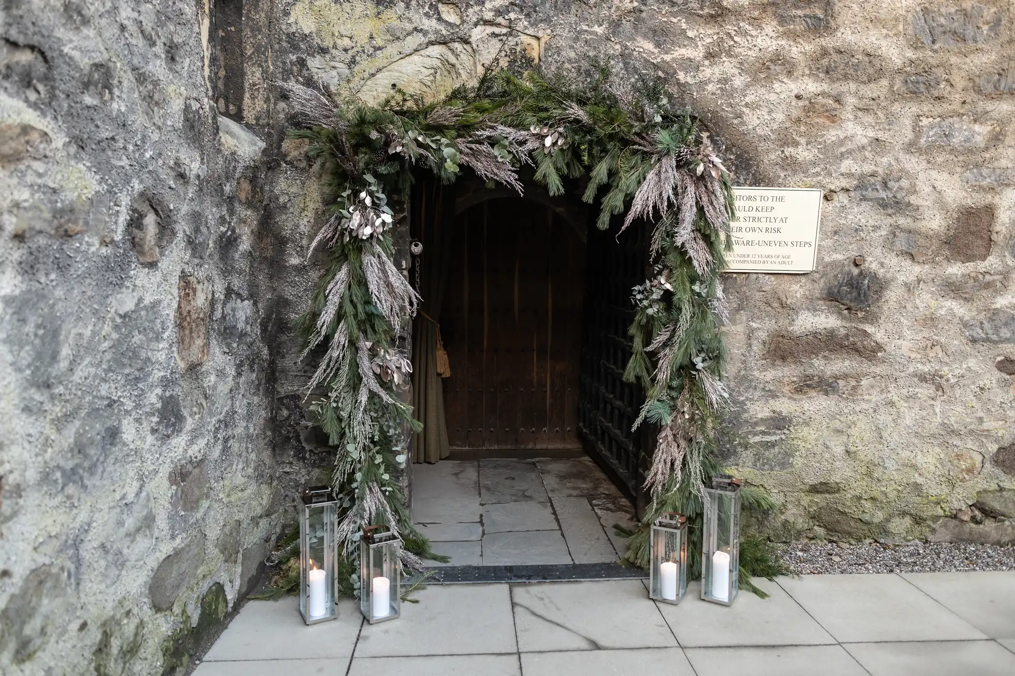 A stone archway adorned with greenery and dried flowers, featuring three lit candles in glass holders at the base. A small sign is visible on the right side of the arch.