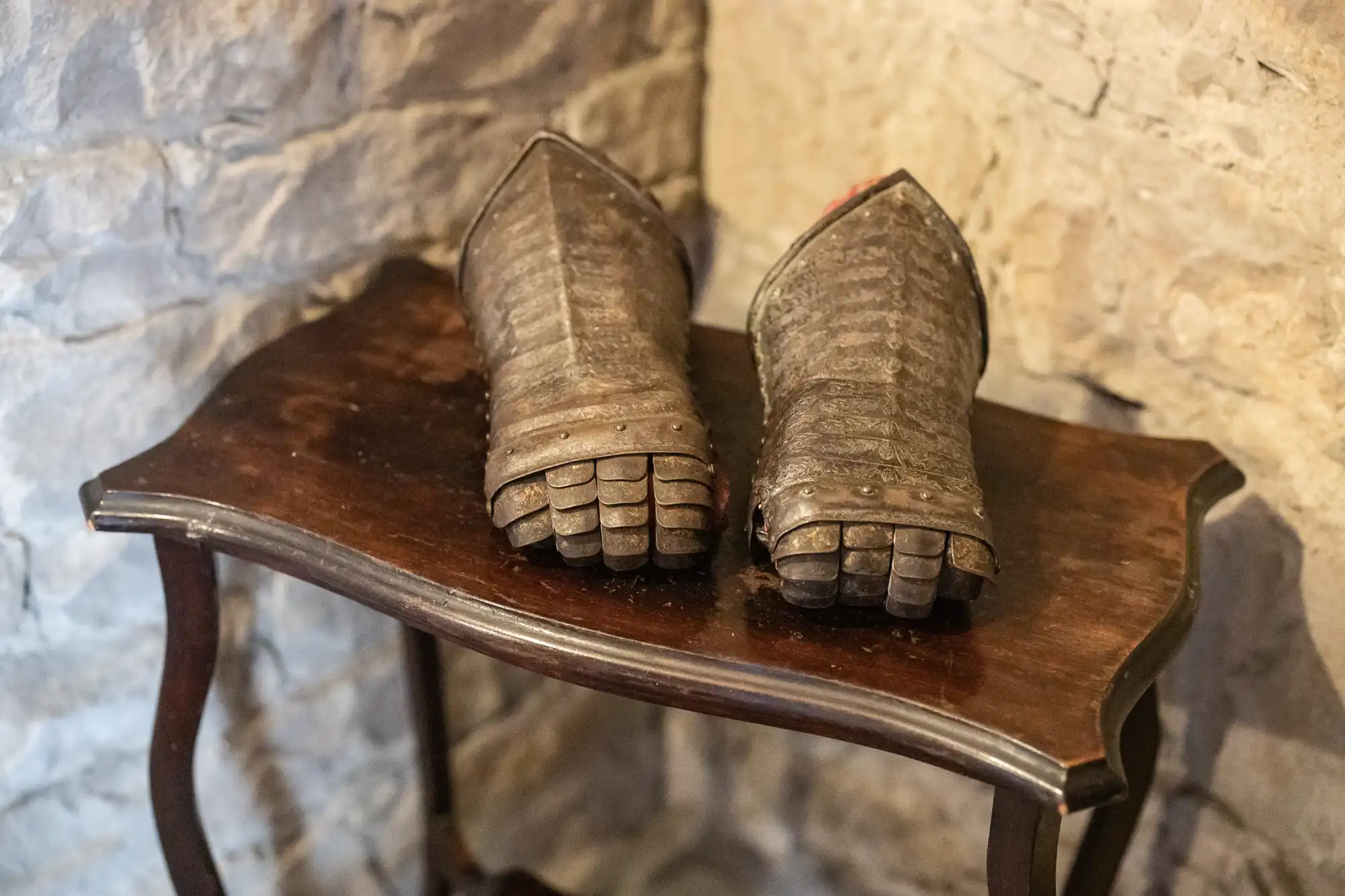 A pair of medieval metal gauntlets with articulated fingers sits on a small wooden table against a stone wall.