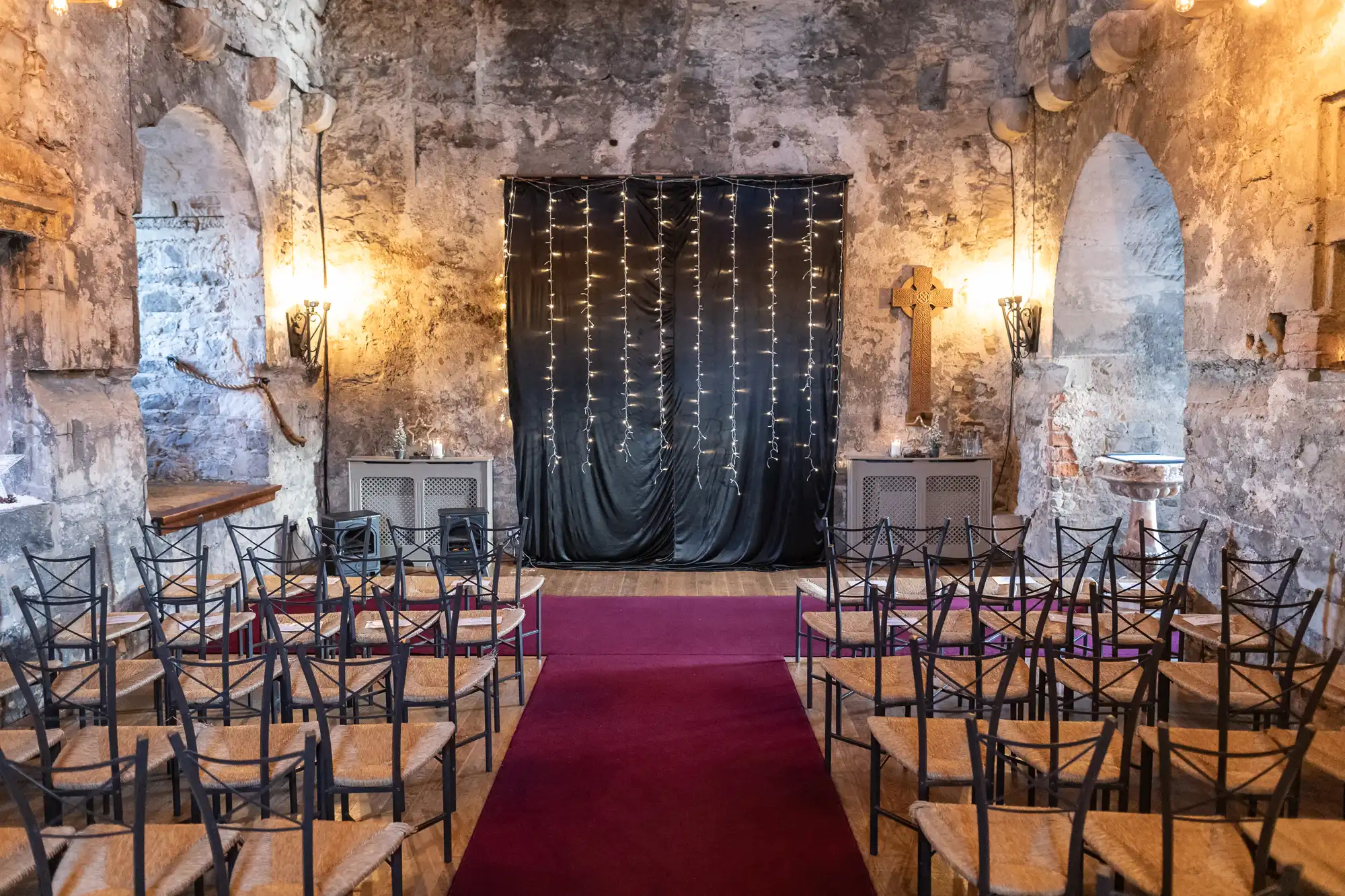 A small wedding venue with stone walls, a red carpet aisle, and rows of chairs. A backdrop with string lights and black fabric is set up at the front, and the space is illuminated by wall sconces.