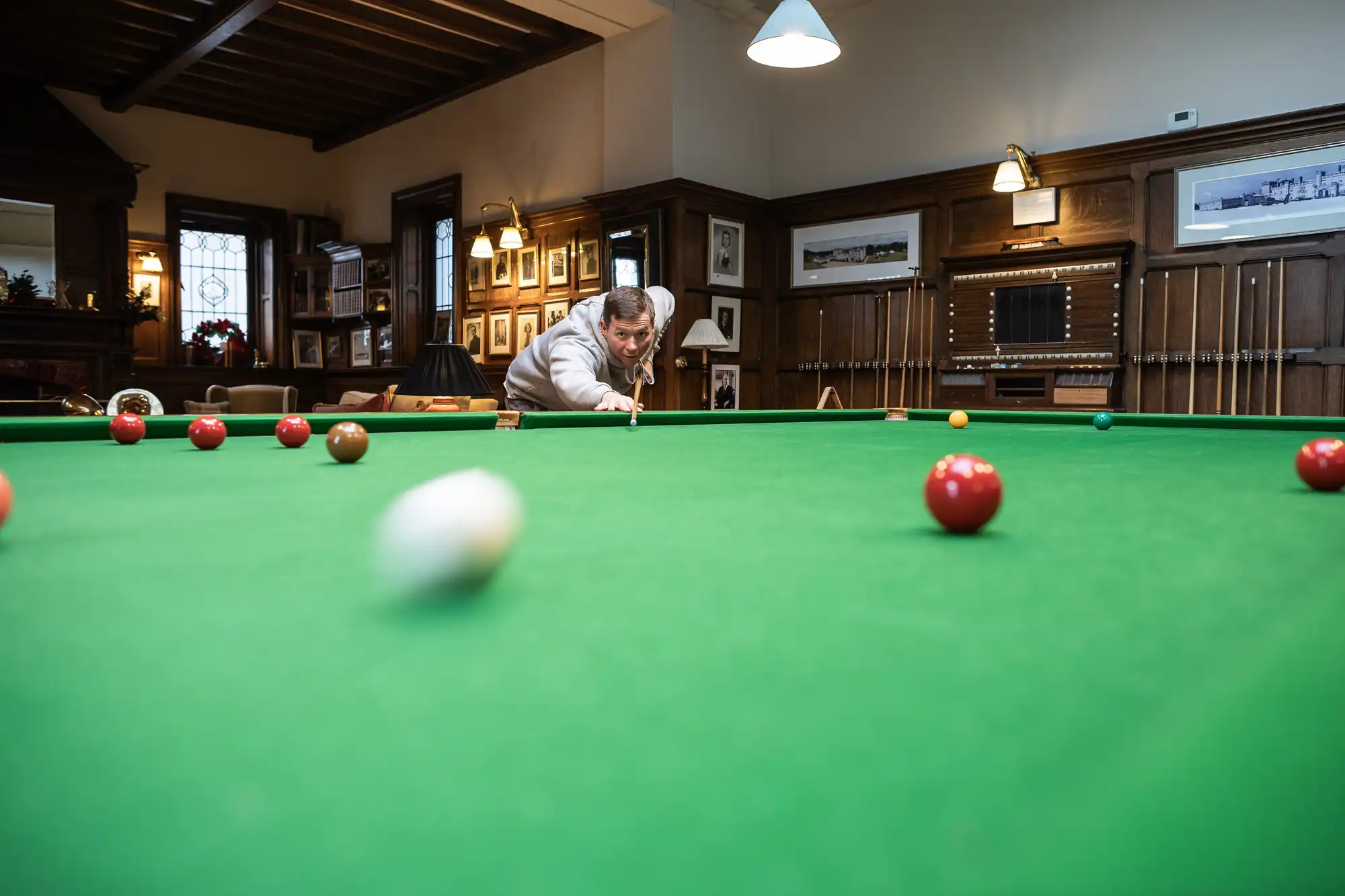 A person is aiming to hit the cue ball on a green pool table, with other balls scattered across the table. The room has wood-paneled walls, framed photos, and multiple pool cues on the rack.