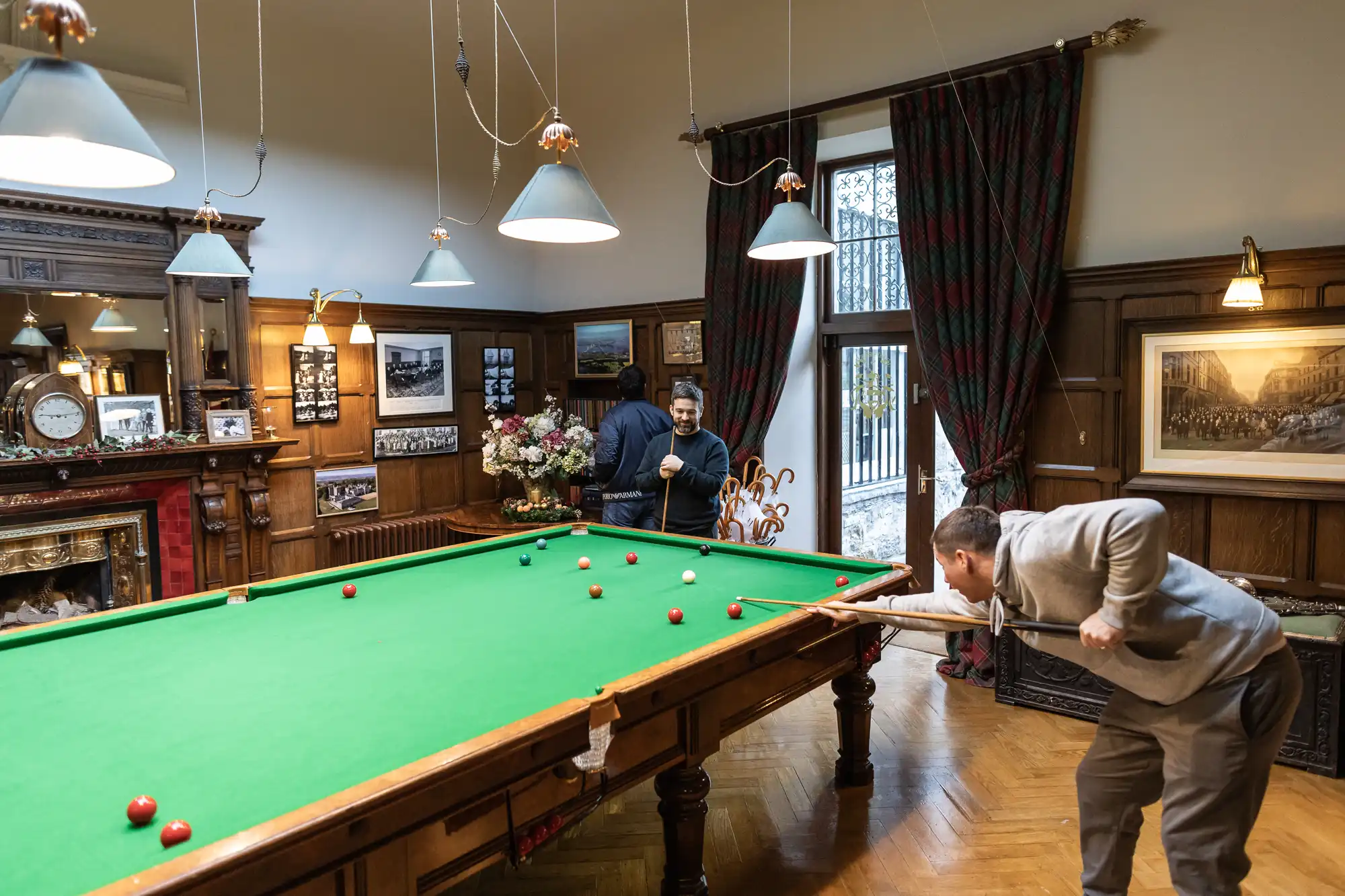 Two people are playing snooker in a richly decorated room with wood-paneled walls, framed photos, and hanging lights. One is taking a shot while the other looks on with a cue stick.