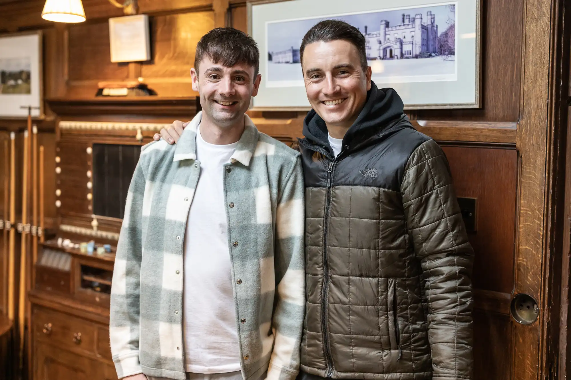 Two men stand side by side in a warmly-lit, wooden-paneled room, smiling at the camera. One wears a white and green plaid jacket, and the other wears a dark green puffer jacket.