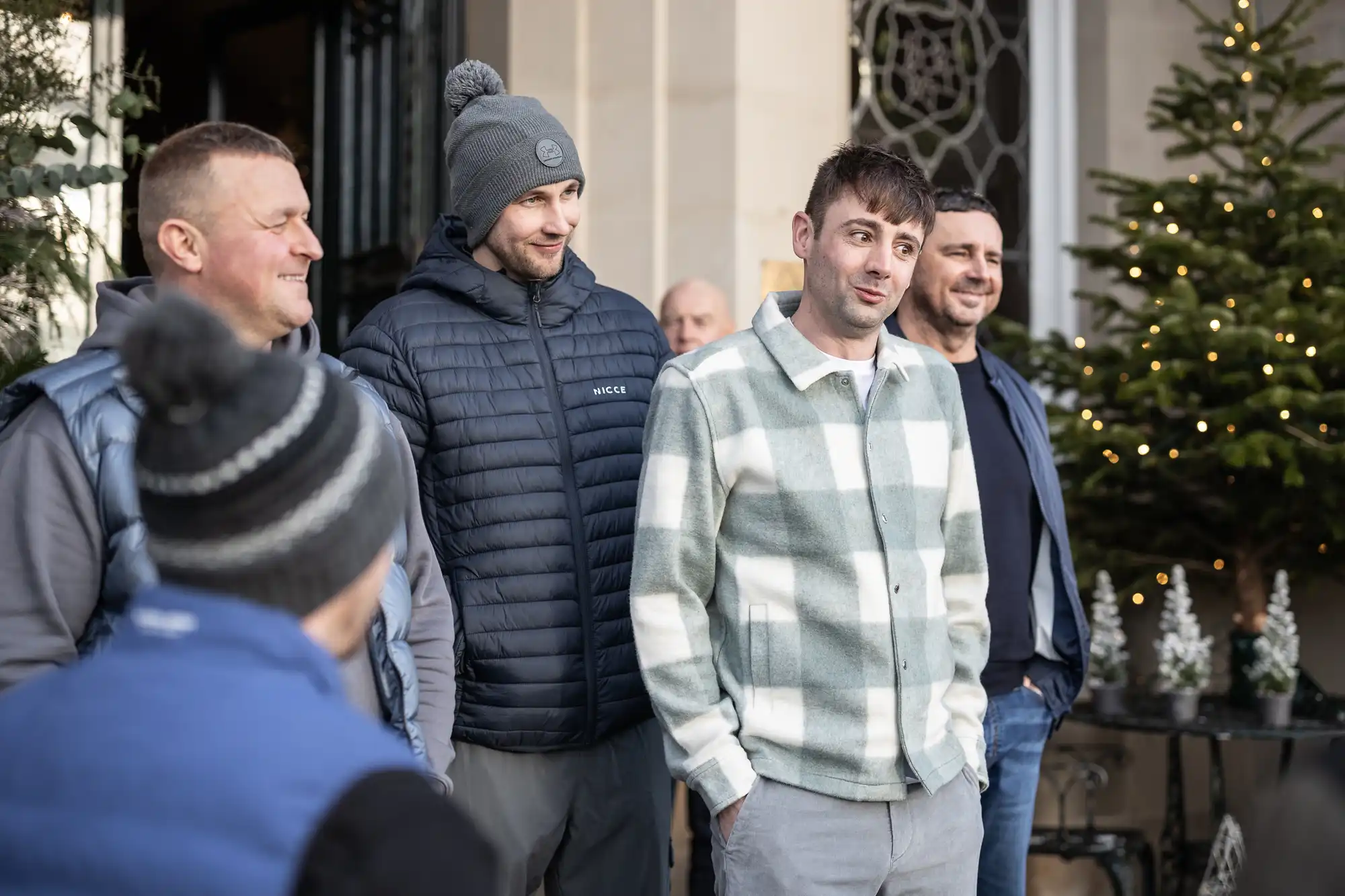 A group of men dressed in casual winter clothing stand outdoors in front of a building with decorated Christmas trees.