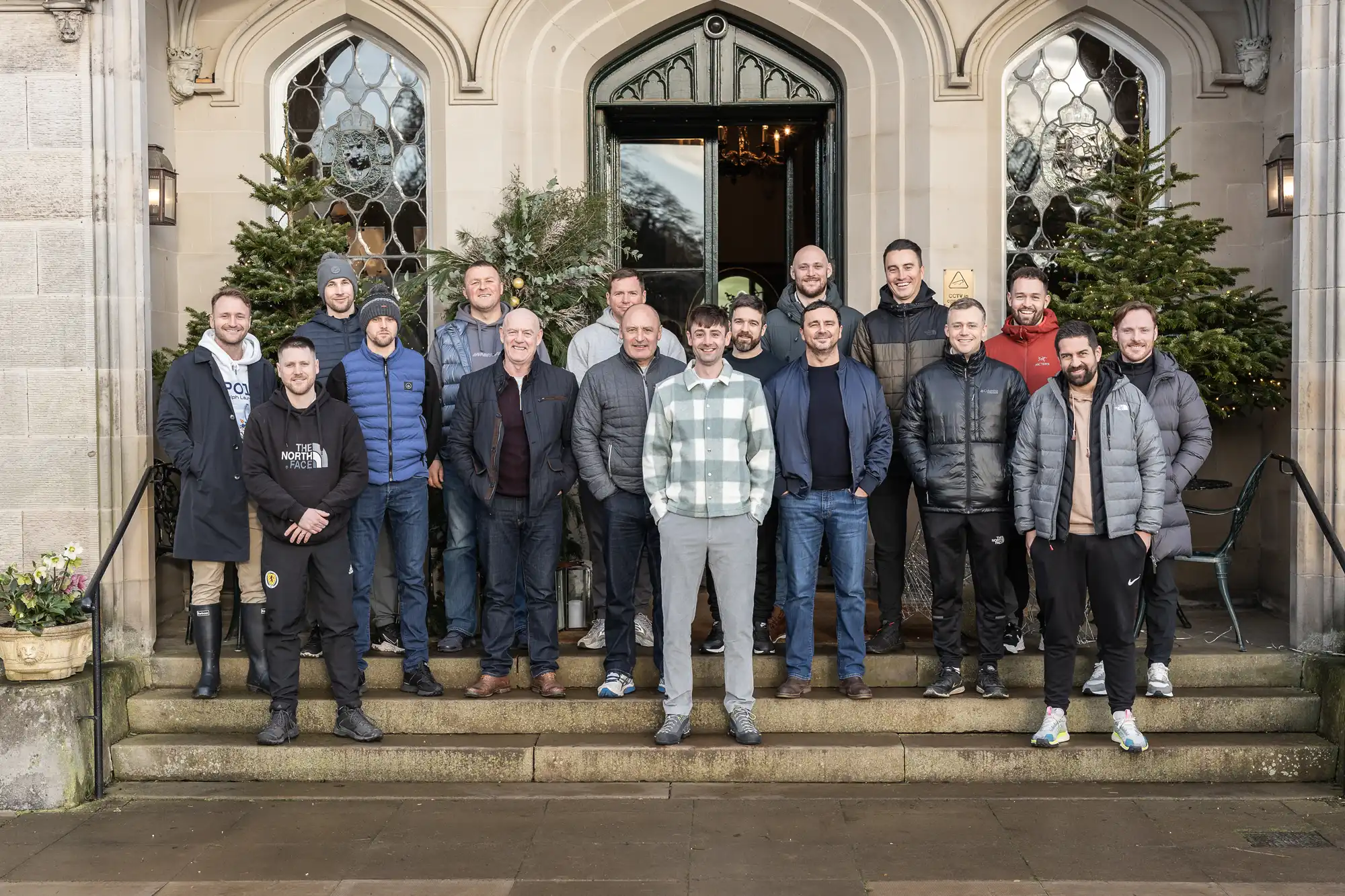 A group of fifteen men stand on the steps of a stone building adorned with two Christmas trees at its entrance. They are casually dressed, with some smiling at the camera.