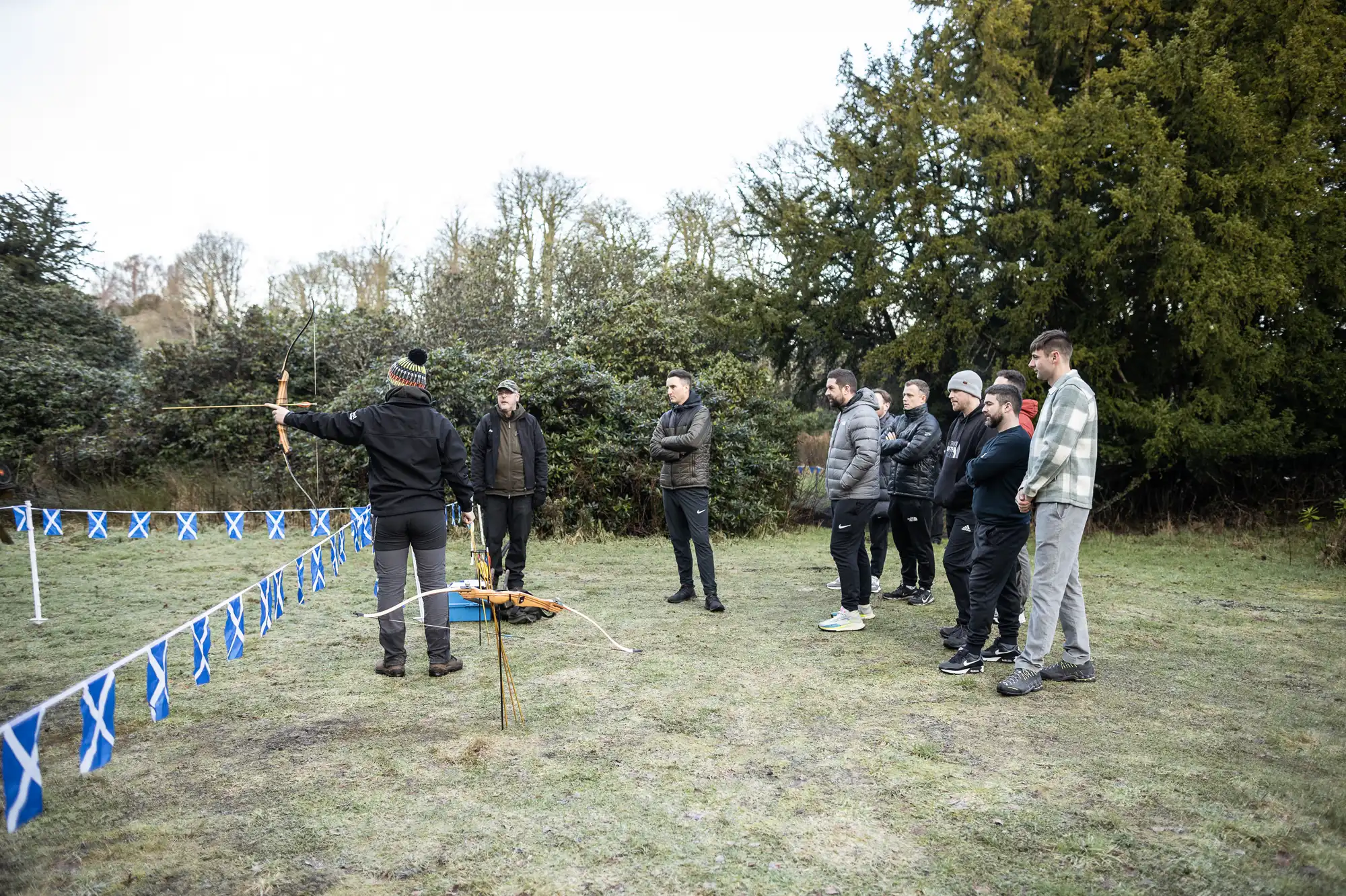 A group of people observes an archery demonstration outdoors. The instructor aims an arrow at a distant target while standing behind a blue and white flagged barrier.