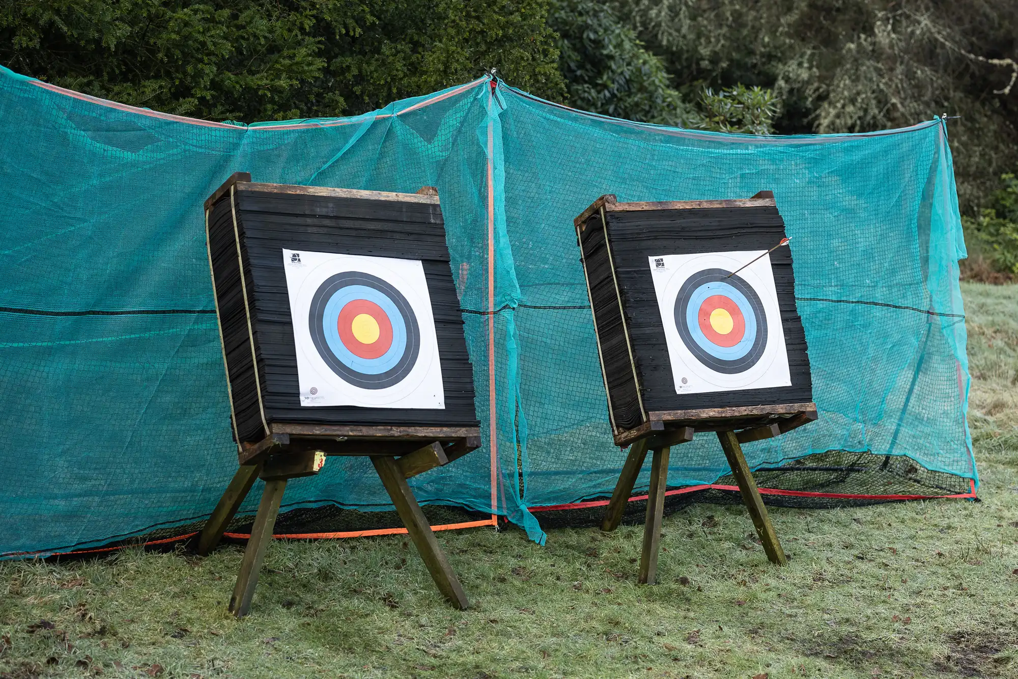 Two archery targets with colorful bullseyes are set up outdoors in front of green netting. An arrow is stuck in the right target near the center. The ground is covered with short grass.