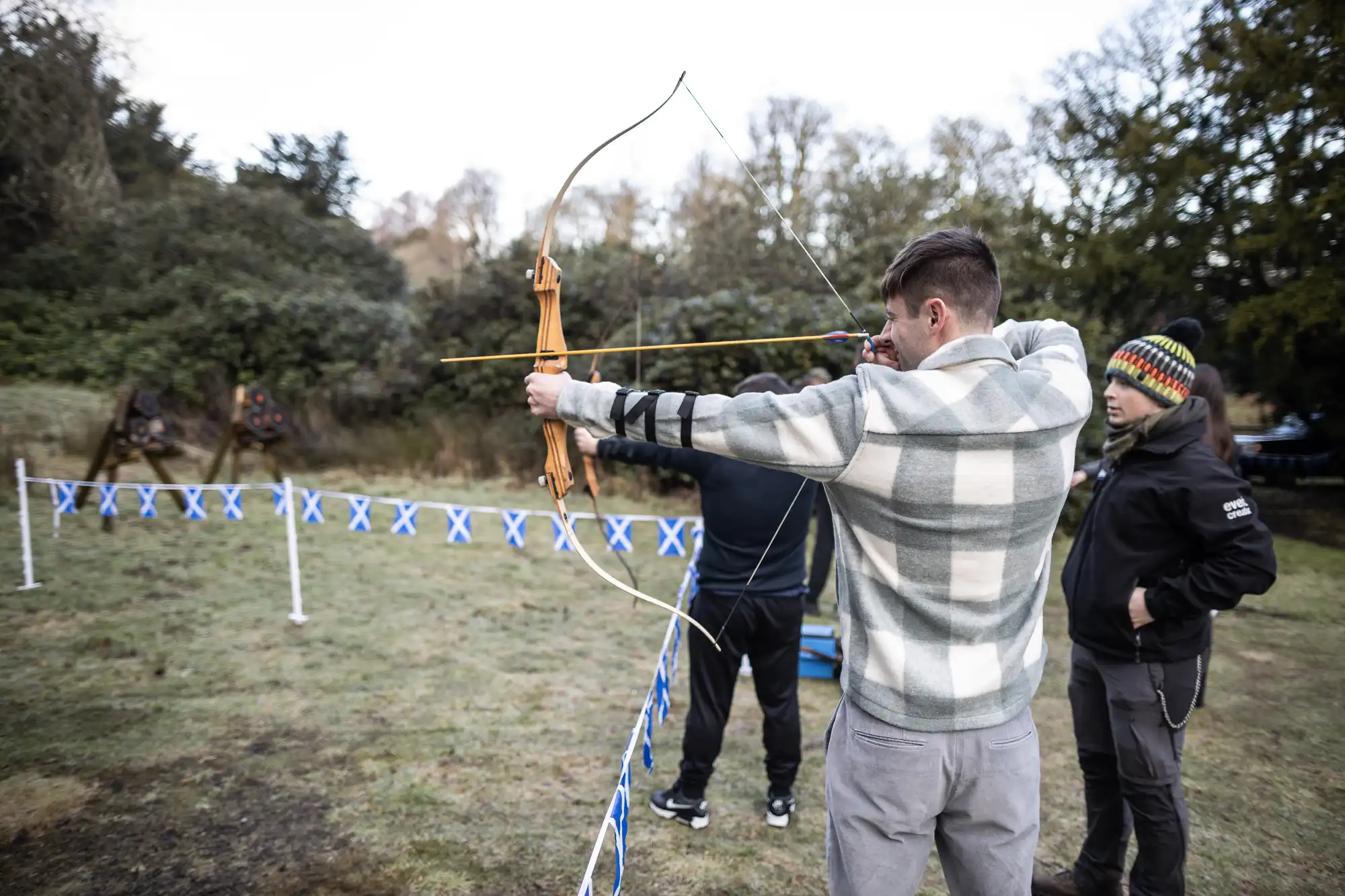 A man in a plaid jacket aims a bow and arrow outdoors, while two others stand nearby, one in a black jacket and colorful beanie. Trees are in the background with a ground marked by blue and white tape.