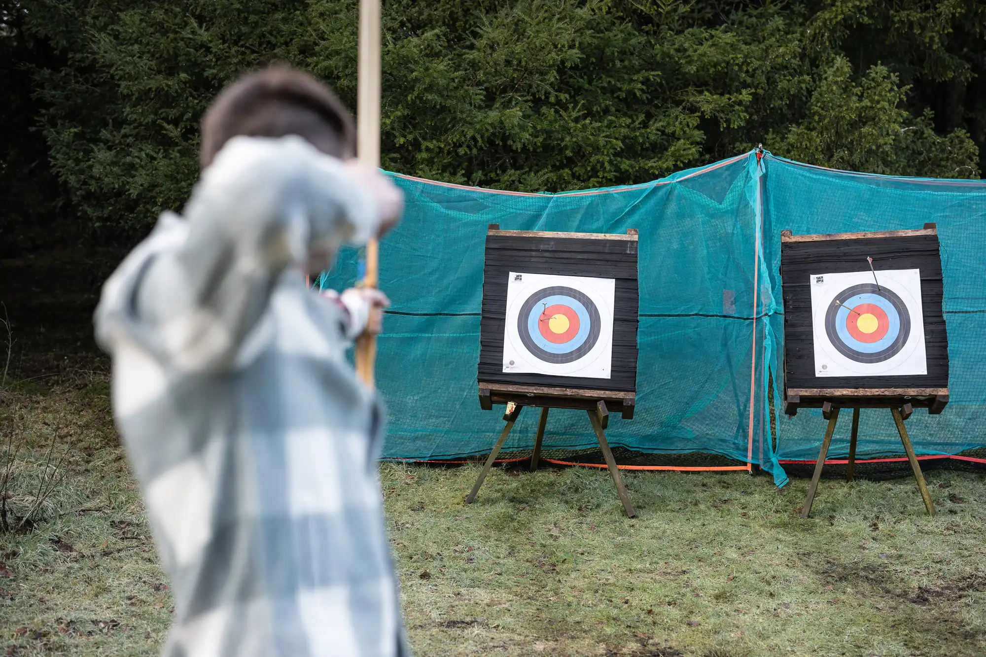 Person aiming a bow and arrow at one of two target boards set up outdoors.