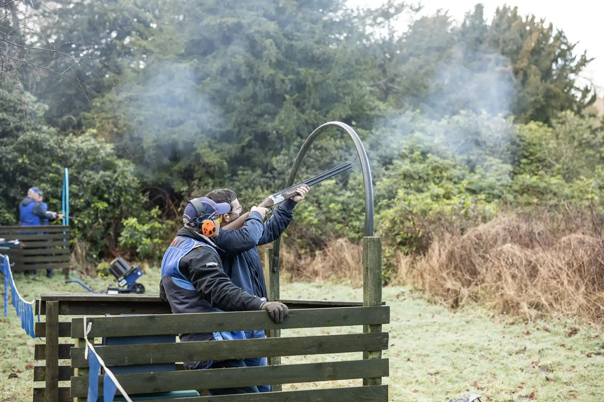 Two individuals in protective gear participate in a clay pigeon shooting activity outdoors, aiming shotguns towards the sky with smoke visible.