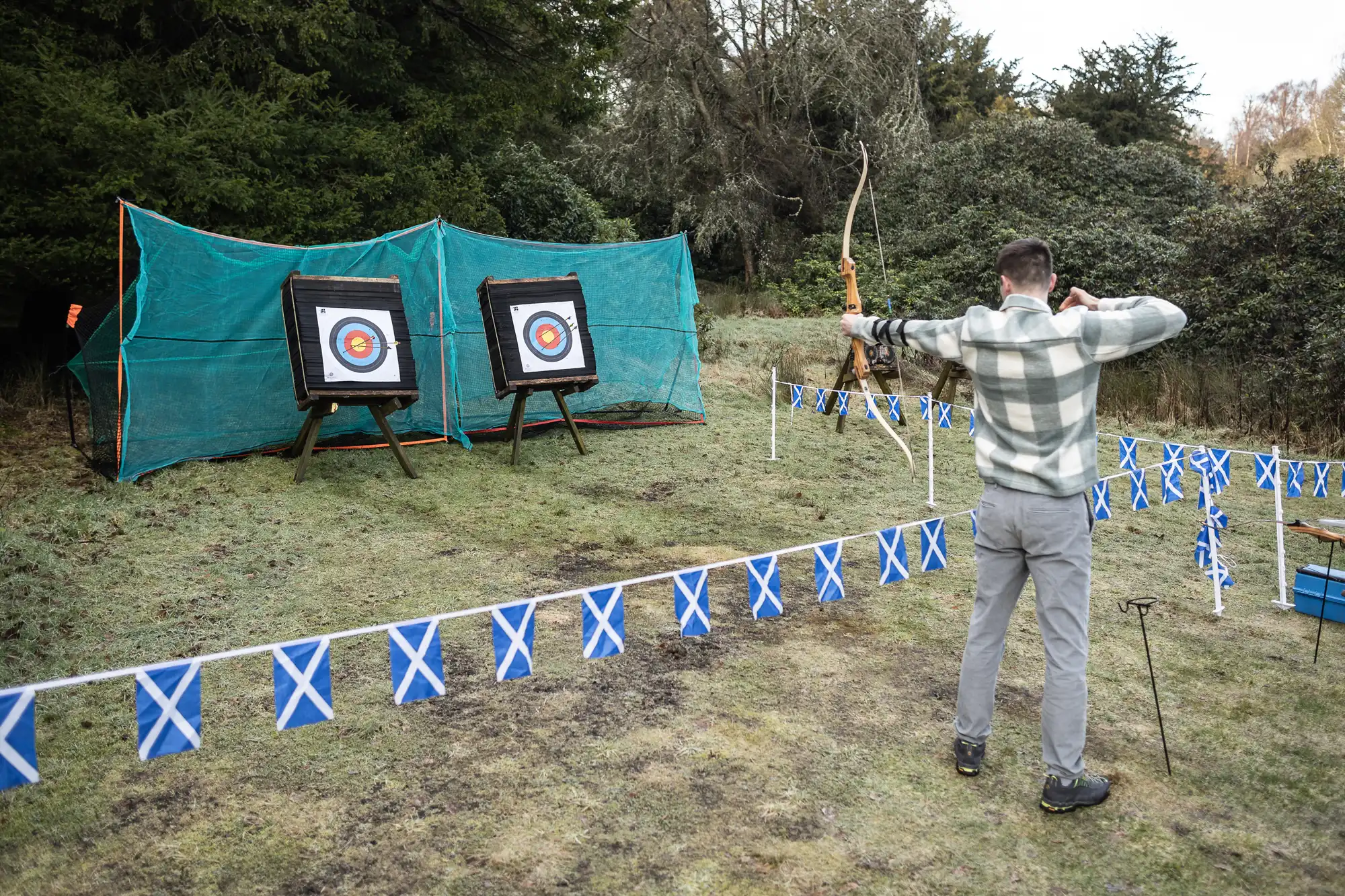 A man in a plaid jacket aims a bow and arrow at two archery targets in an outdoor setting. The area is fenced off with blue and white flag bunting.