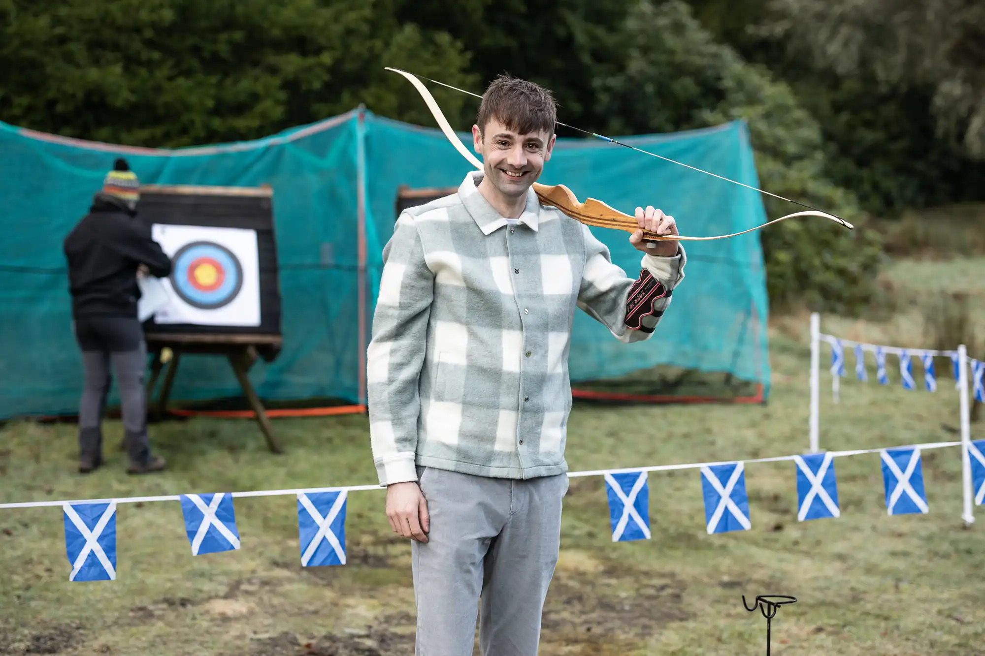 A person stands holding a bow with an archery target in the background. Another individual is adjusting the target. Blue and white flags are visible in the foreground.