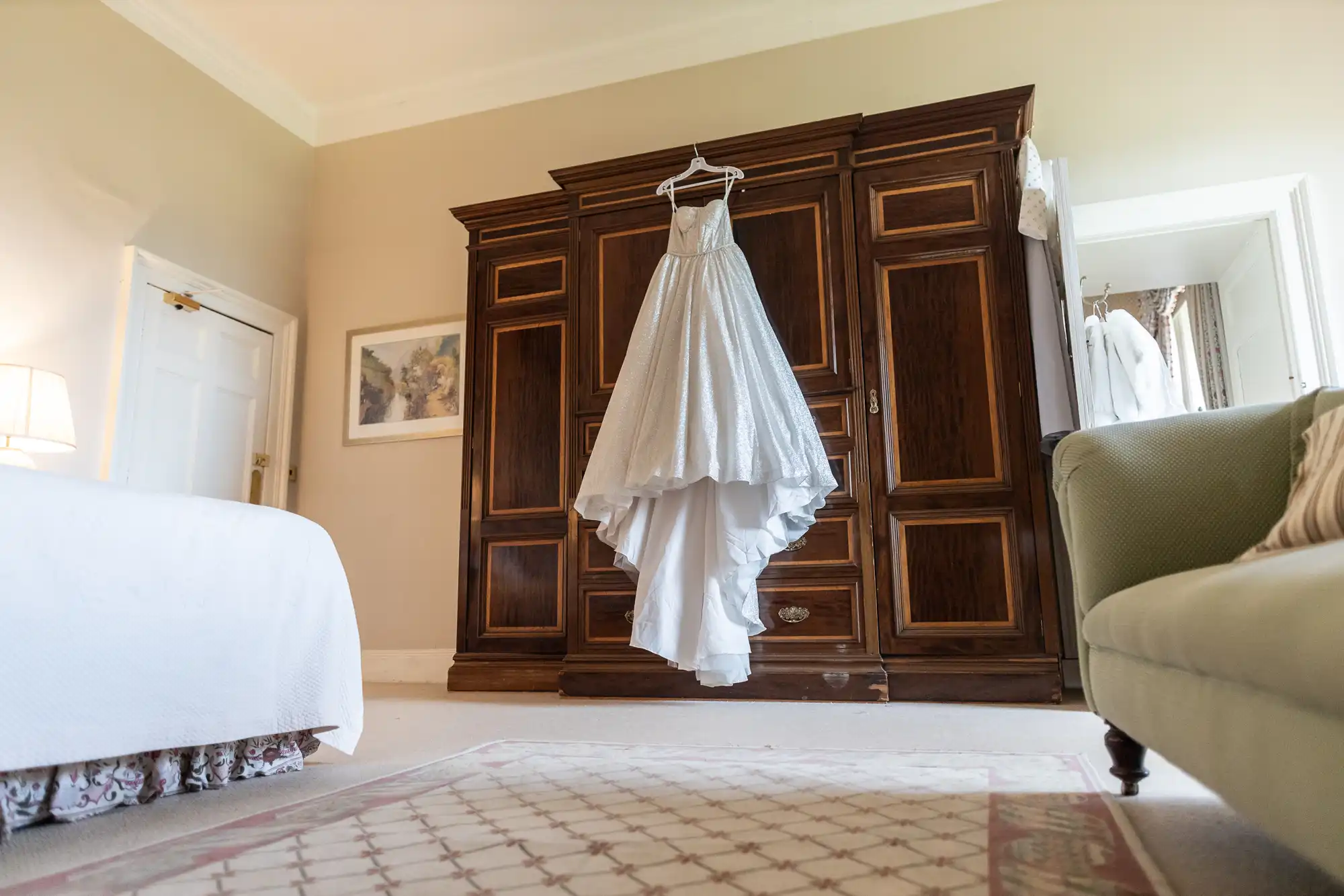 A white wedding dress hangs on a wooden wardrobe in a well-lit bedroom with beige walls, a green couch, and a bed covered in white linens.