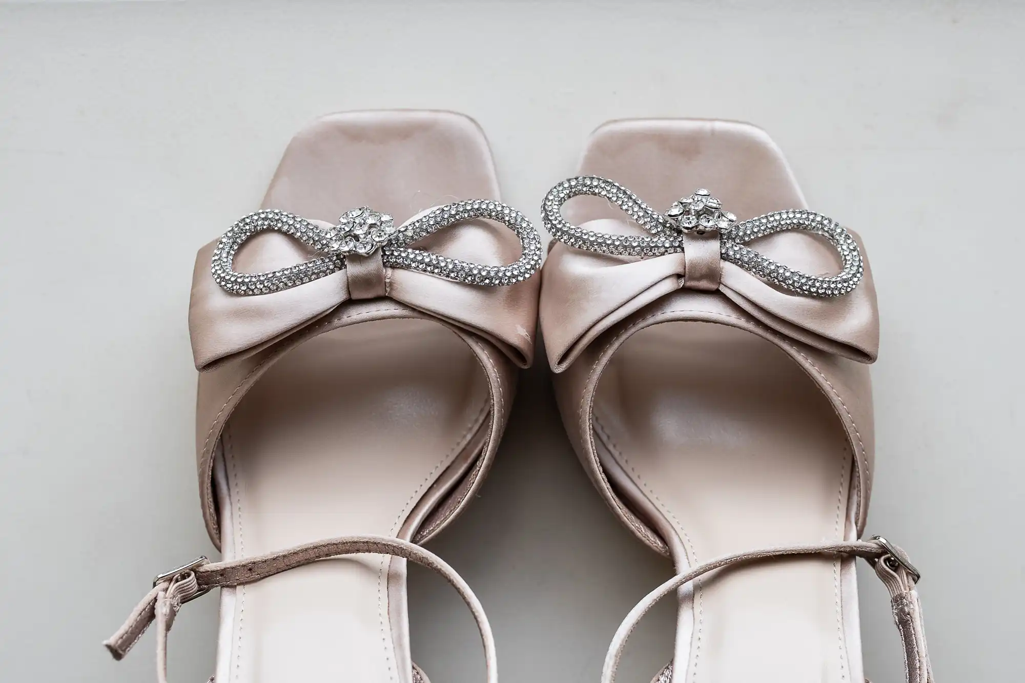 A pair of pale pink high-heeled open-toe shoes with bow and rhinestone embellishments on a plain background.