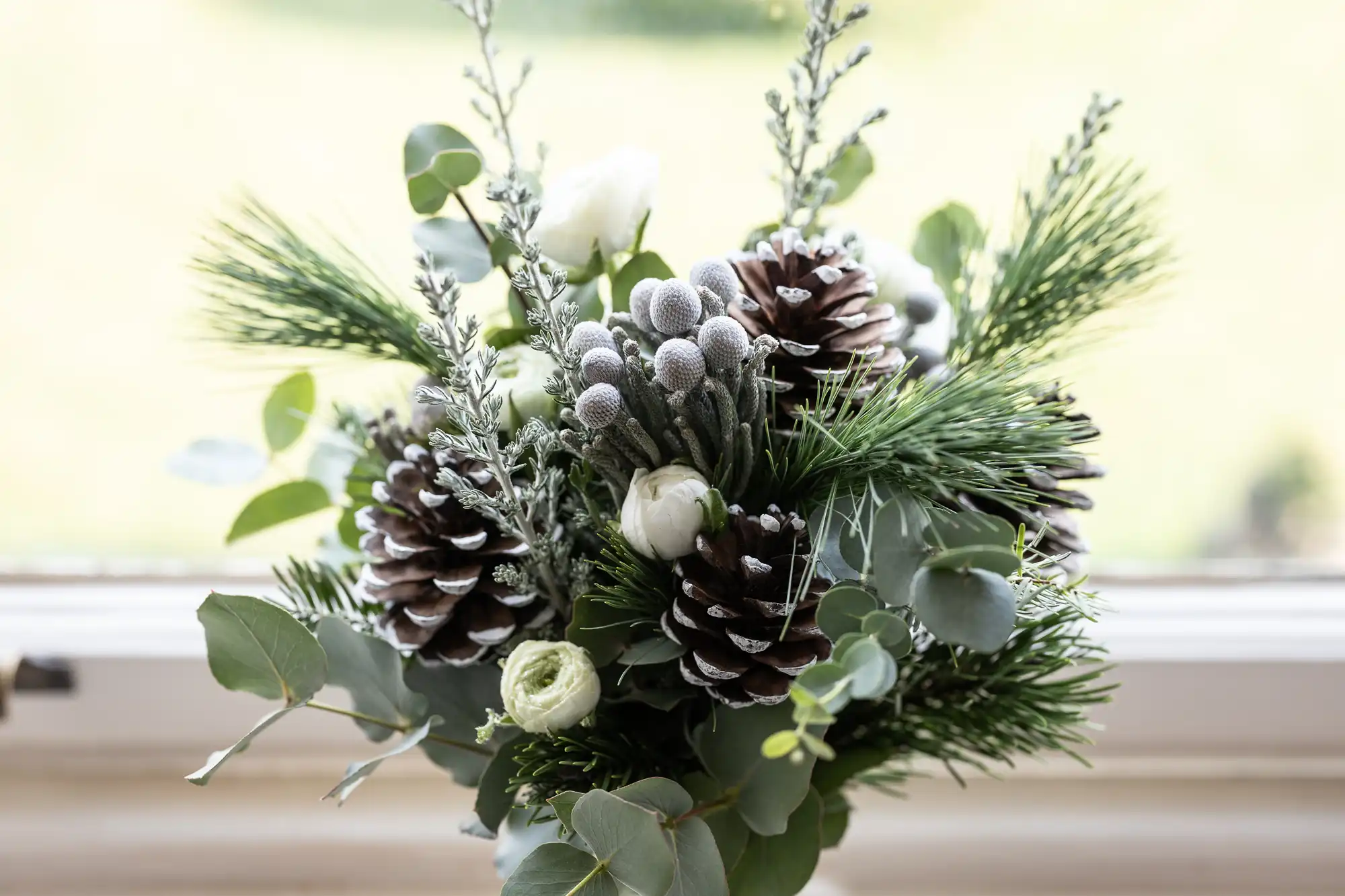 A flower arrangement with pine cones, green foliage, and white flowers is placed on a window sill.