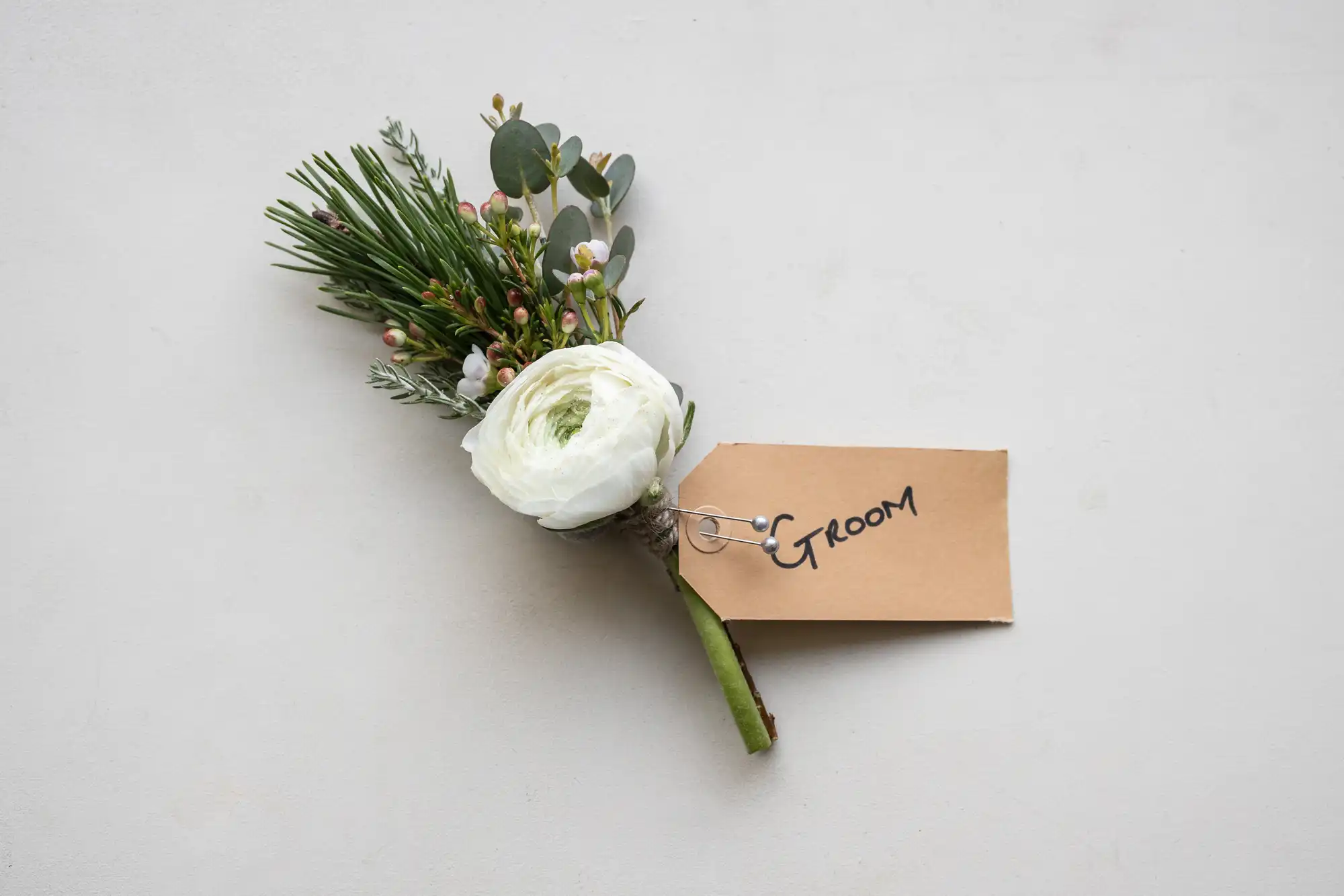 A boutonniere with a white flower, greenery, and small berries attached to a beige tag labeled "GROOM" on a white background.