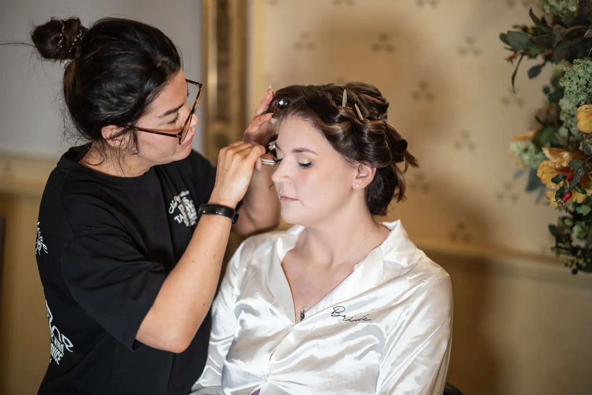 A makeup artist applies eyeshadow to a seated woman with rollers in her hair, who is wearing a white satin robe.