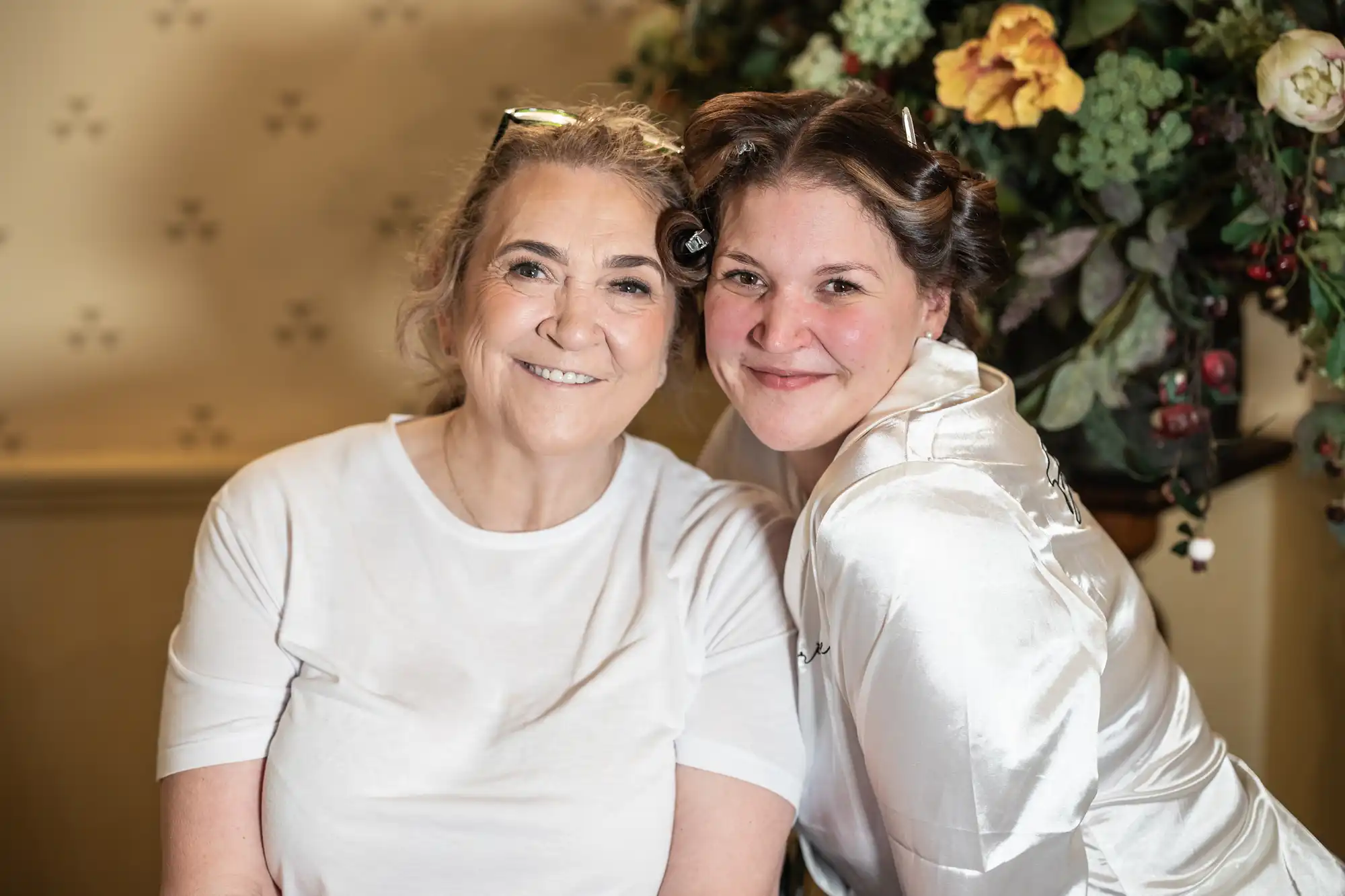 Two women smiling indoors, one in a white T-shirt and the other in a white satin top with curlers in her hair, standing close to each other in front of a floral arrangement.