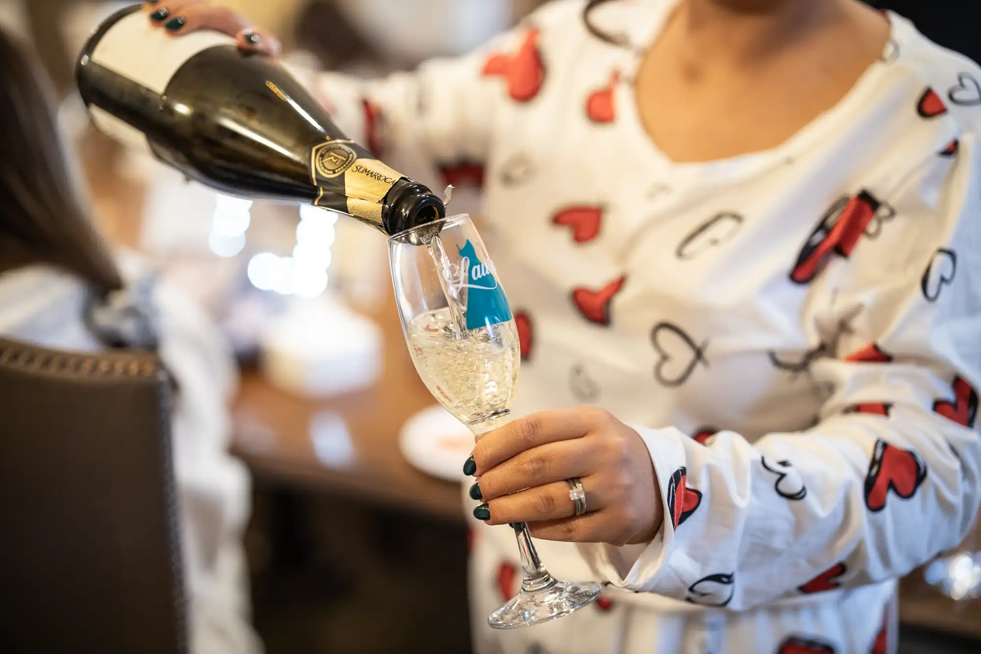 A person wearing a white shirt with red and black heart patterns pours champagne into a glass.