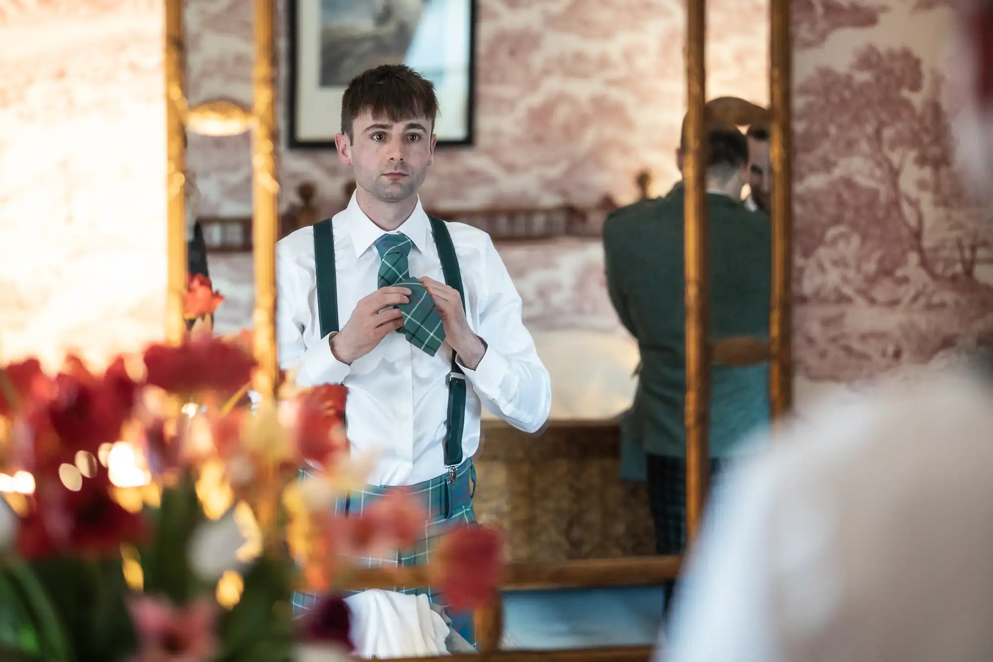 A man in a white shirt and plaid kilt adjusts his tie in a mirror, with a floral arrangement in the foreground.