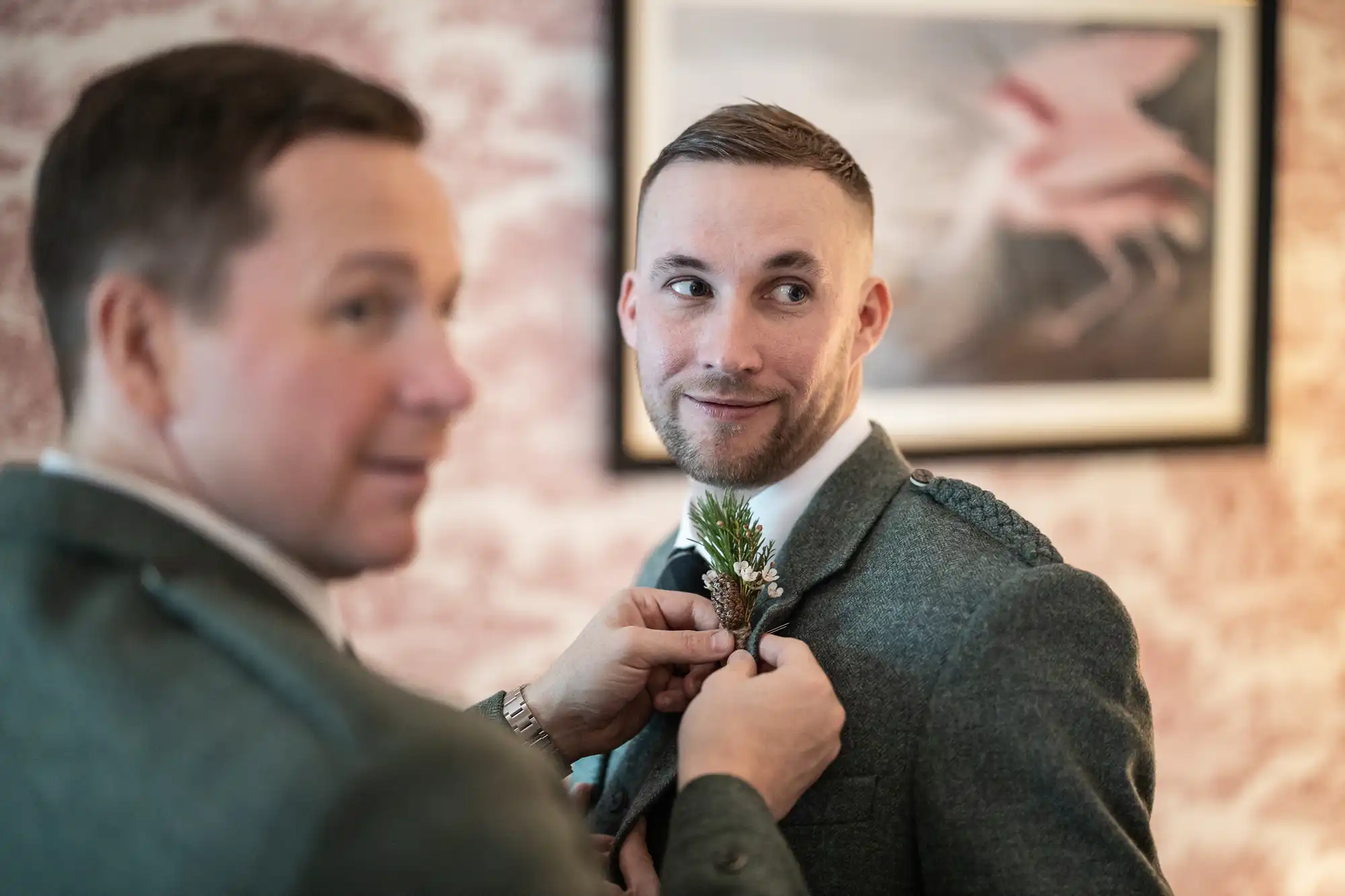 Two men in suits, one of them fastening a boutonniere to the other's lapel. The background features a framed picture and a textured wall.