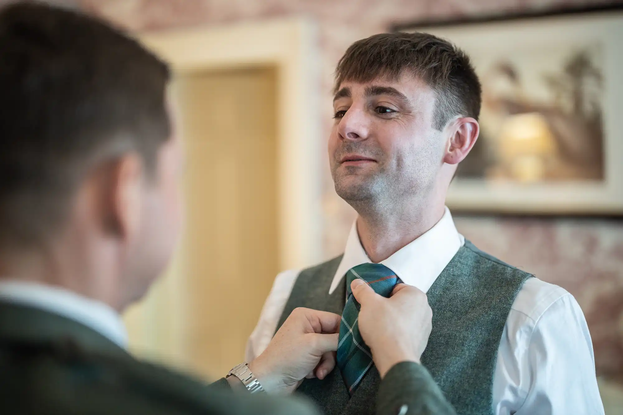 A person is adjusting the tie of another man who is dressed in a gray vest and white shirt. The background features a framed picture on the wall.