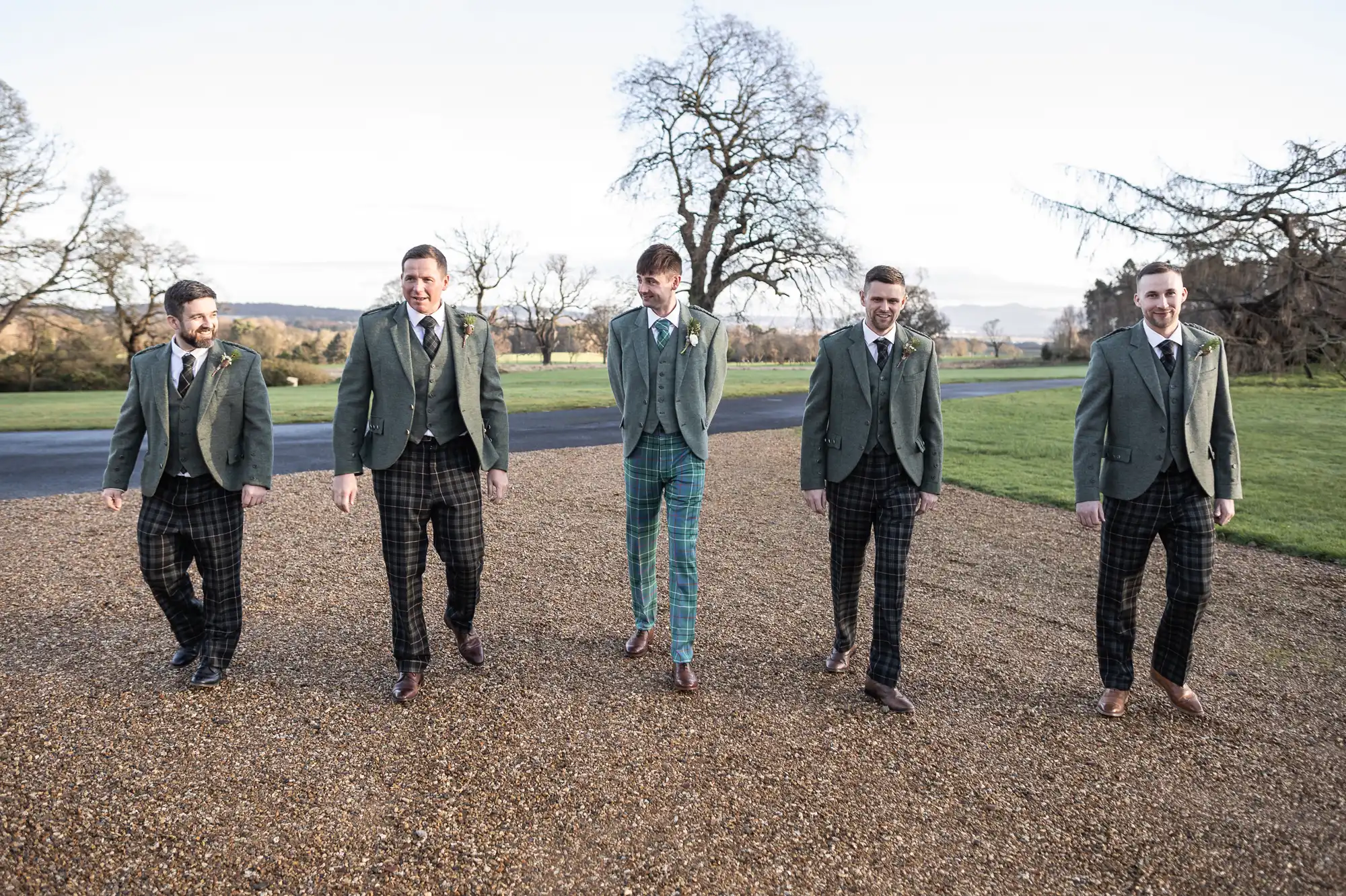 Five men in green jackets with ties and patterned trousers walk outside on a gravel path with trees and grass in the background.