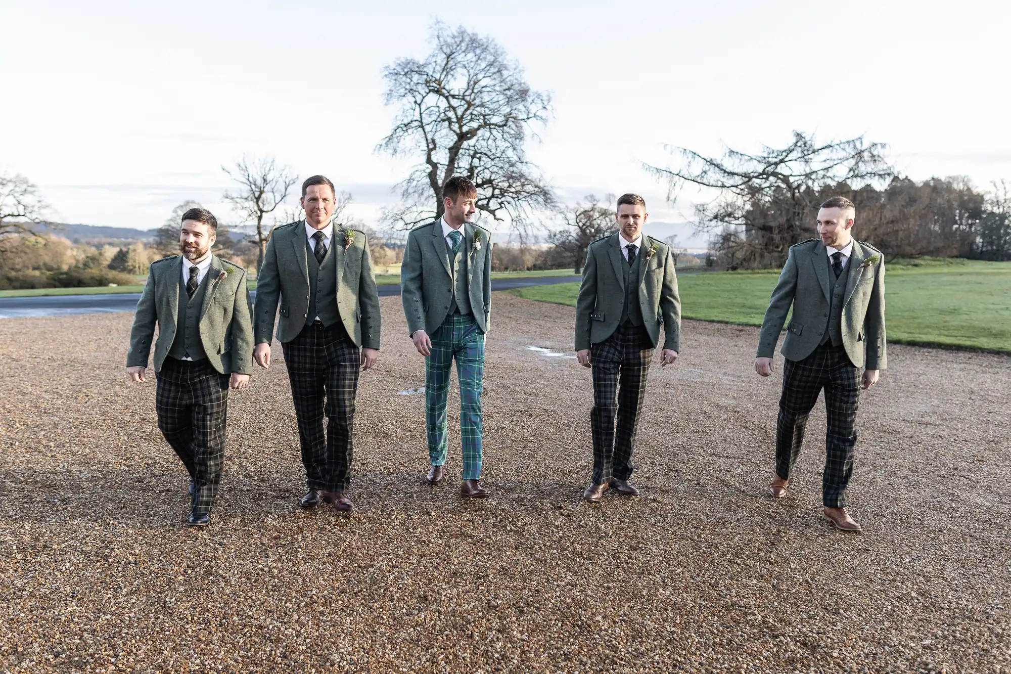 Five men in green blazers and patterned trousers walk together on a gravel path with grassy areas and trees in the background.