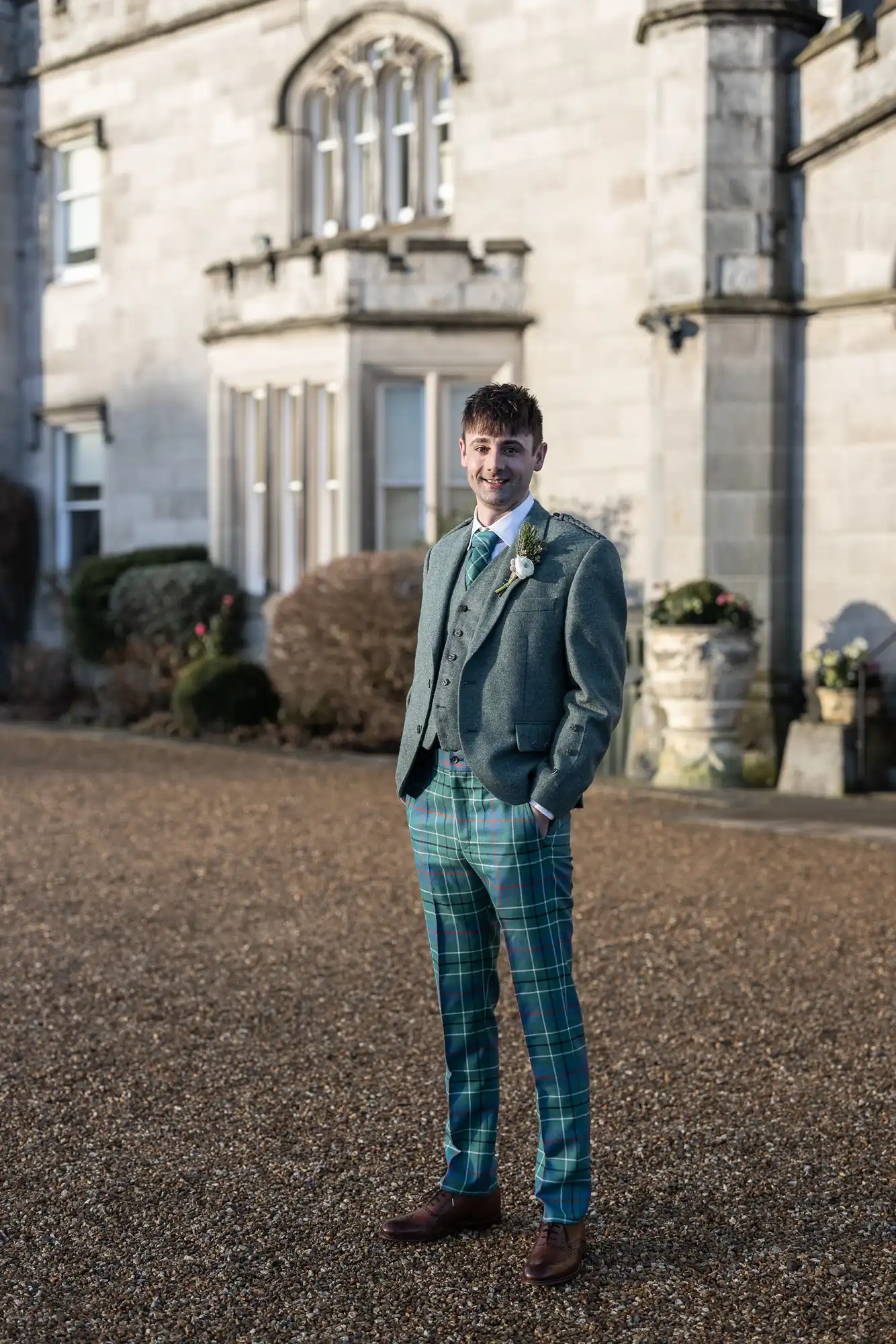 A man wearing a green tweed jacket and green plaid trousers stands outside a stone building on a gravel path.