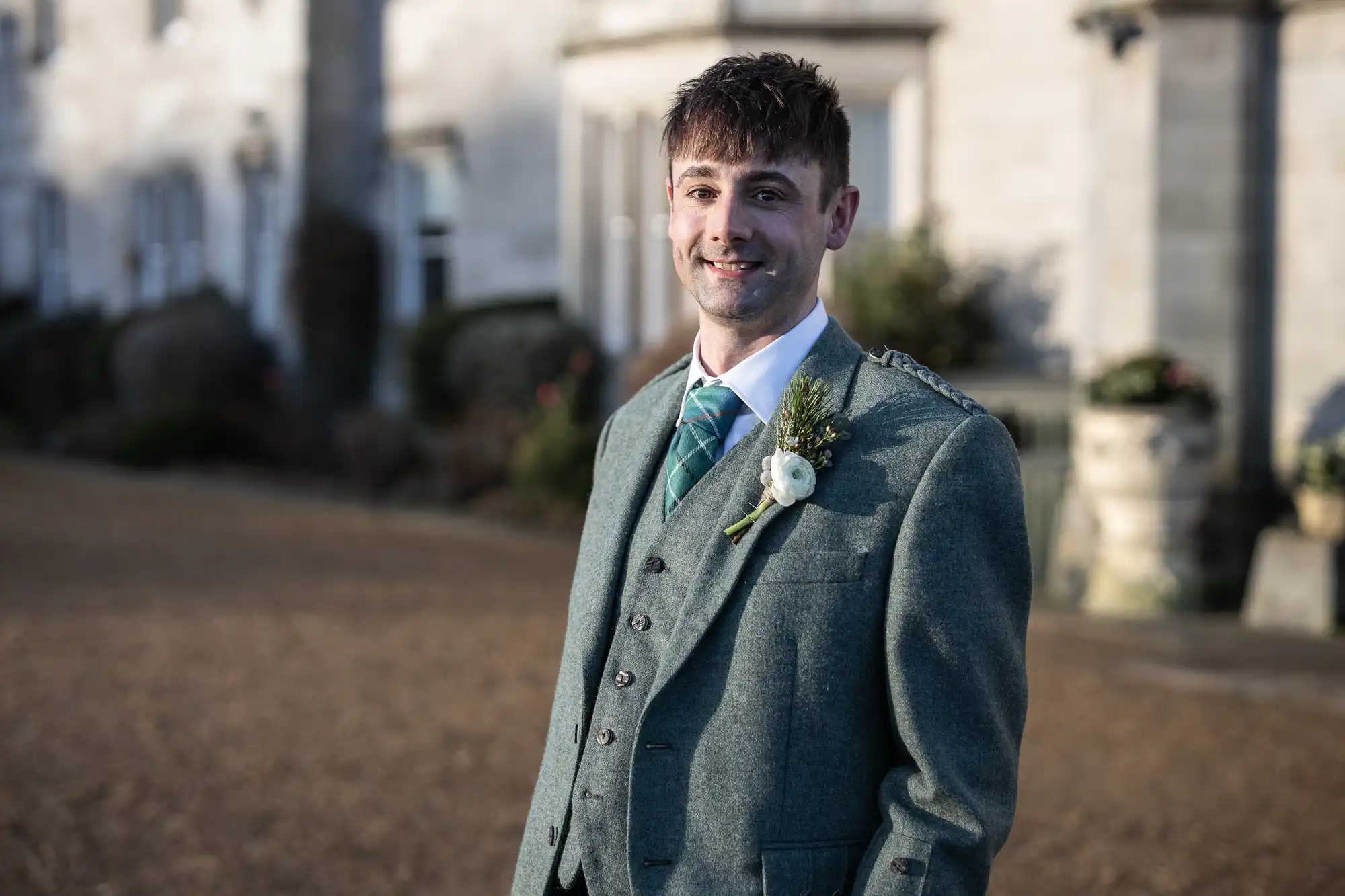 A man in a green suit, wearing a blue tie and boutonniere, stands outdoors in front of a large building.