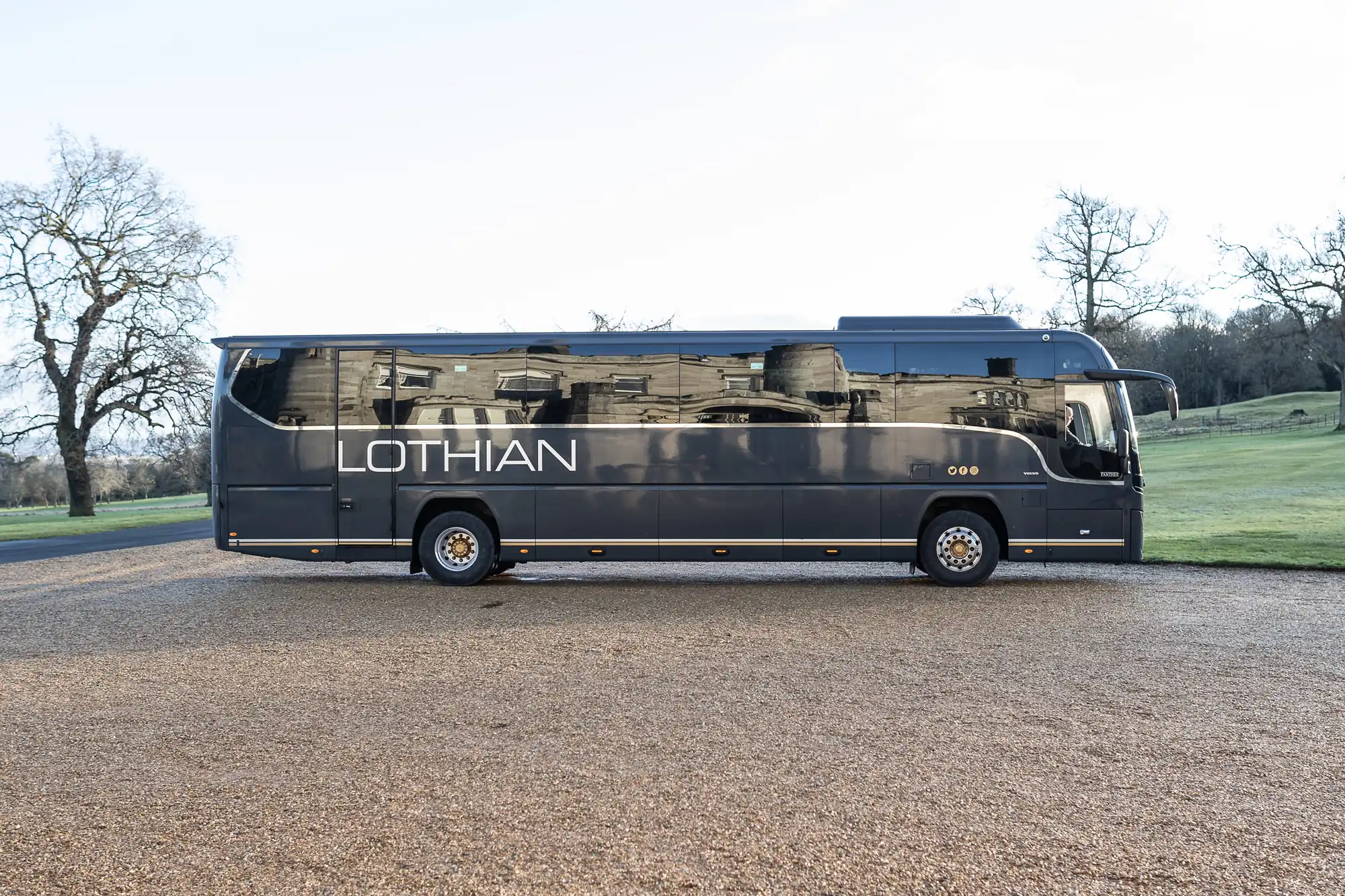 A dark-colored coach bus with the word "Lothian" on its side is parked on a gravel surface with a backdrop of trees and grass.