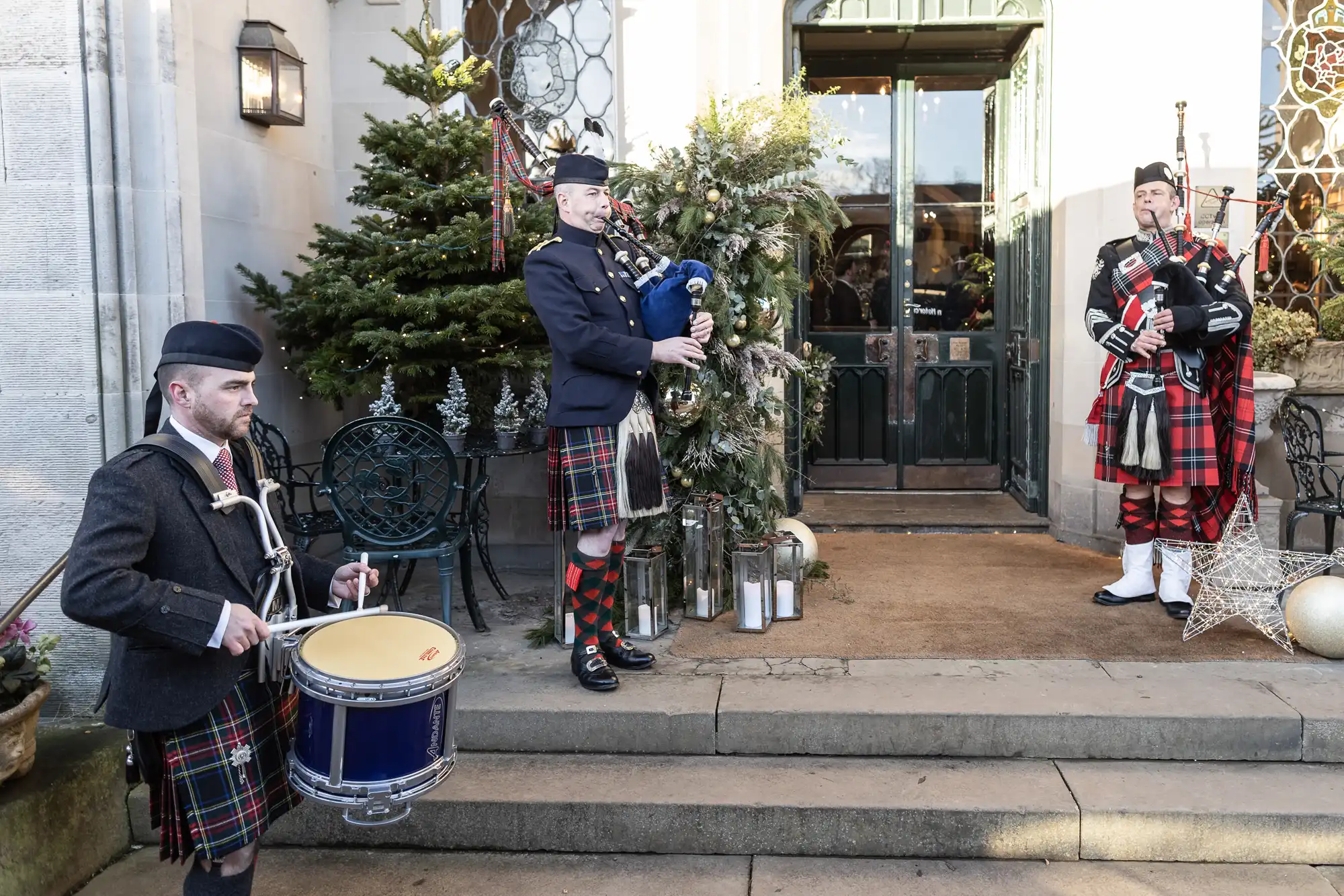 Three men in traditional Scottish attire play musical instruments, including bagpipes and drums, on the steps of a building decorated with greenery.