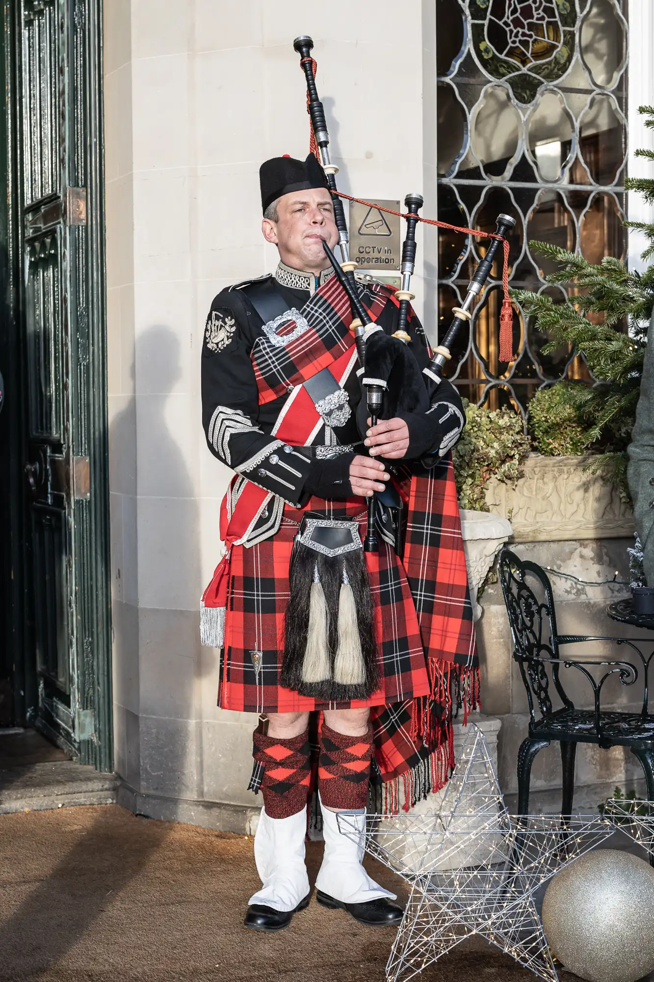 A man in traditional Scottish attire stands playing bagpipes in front of a building adorned with Christmas decorations. He wears a red tartan kilt, black jacket, and white sporran.